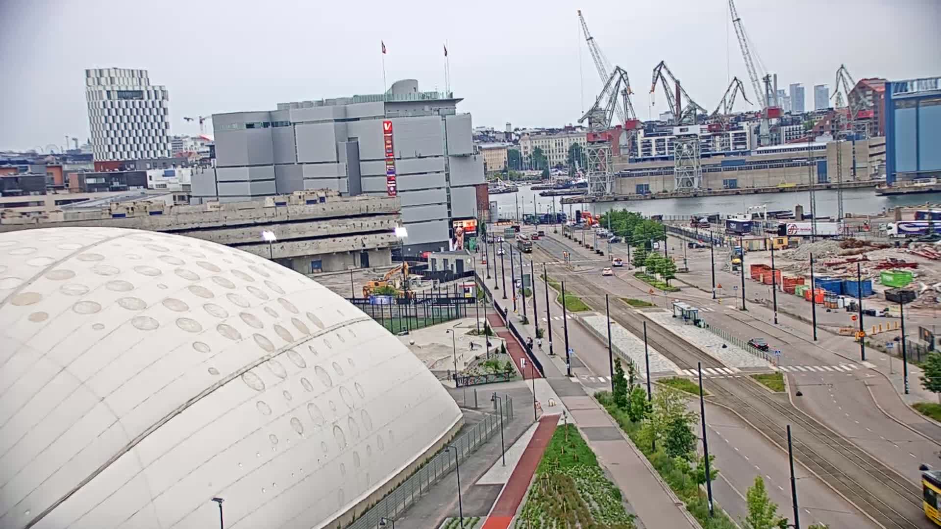 A high-angle view shows a large white domed building, a city street with tram tracks, construction sites, and a harbor with cranes under an overcast sky.