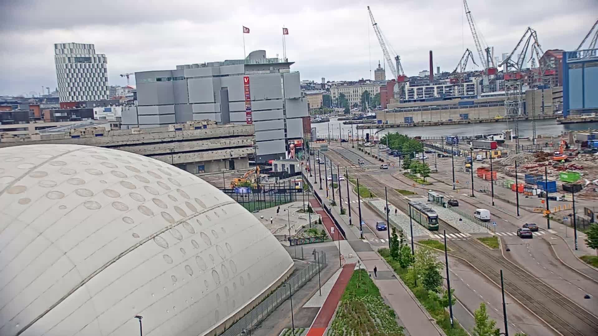 A large domed building sits near a waterfront city scene under an overcast sky, with a tram running along a wide road lined with trees and undergoing some construction.