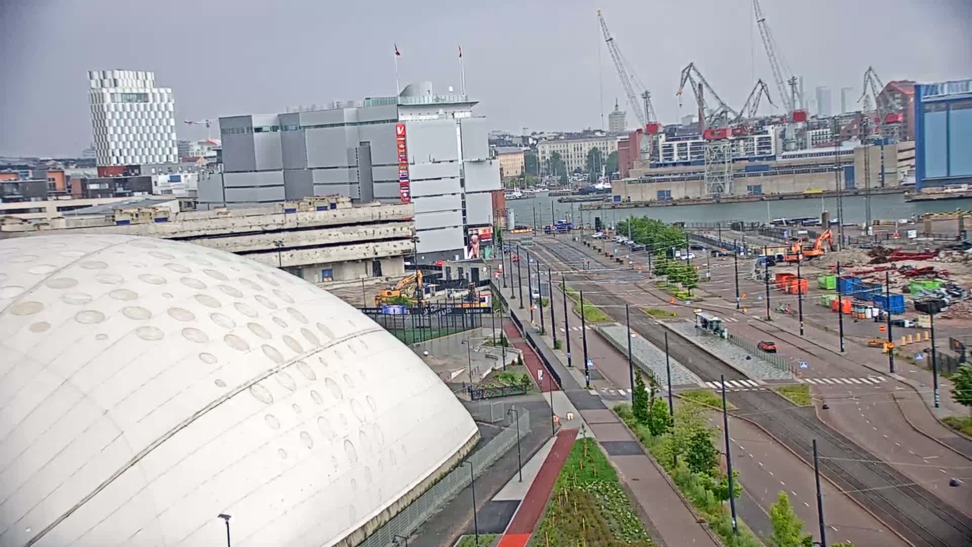A large white dome-shaped structure sits near a waterfront city under an overcast sky, with a street, tram tracks, and construction visible in the foreground.