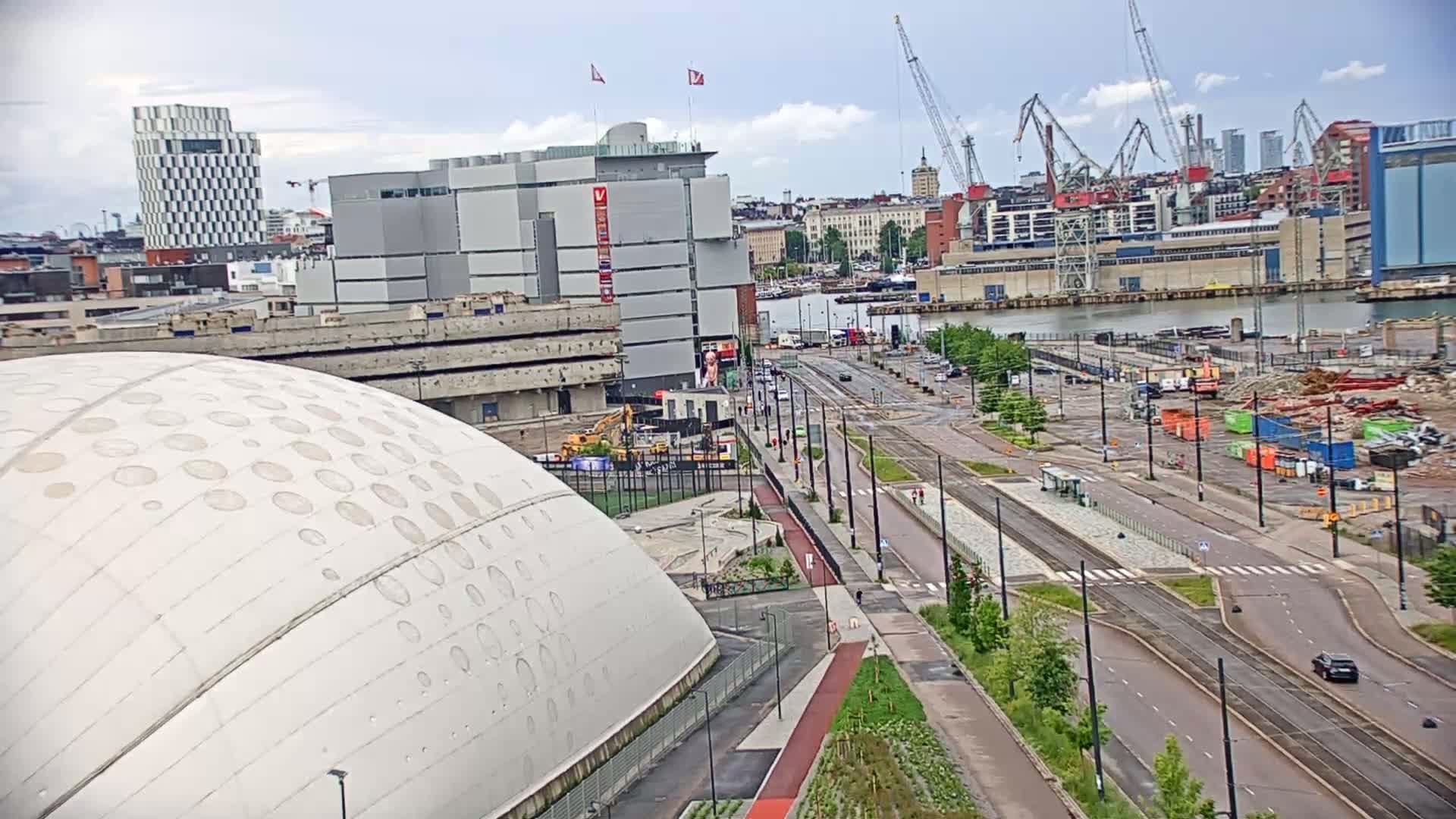 A large domed building is seen next to a wide road with tram tracks running through a city waterfront under cloudy skies.