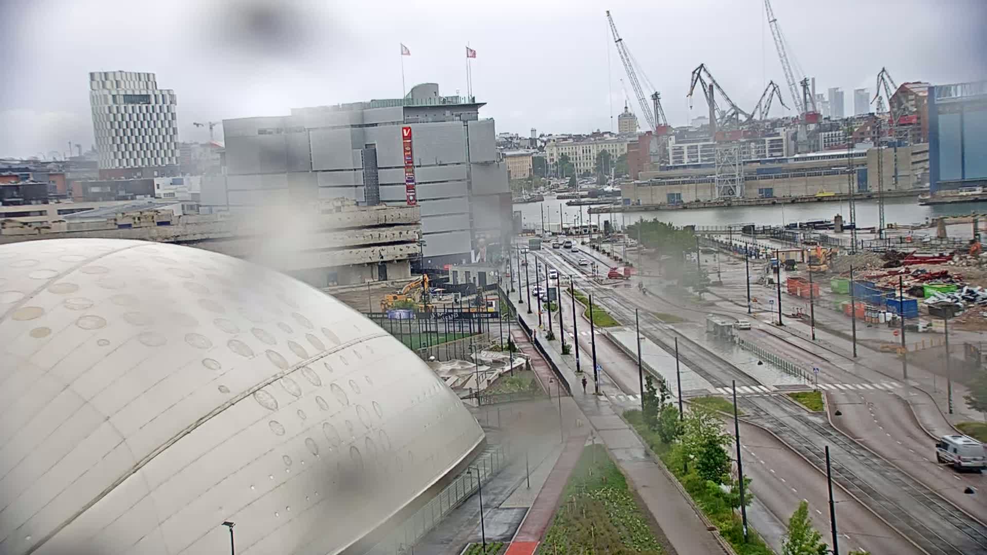 A rain-slicked city street scene shows a large domed building in the foreground, a multi-story building in the mid-ground, a harbor with cranes in the background, and a construction site visible across the street.