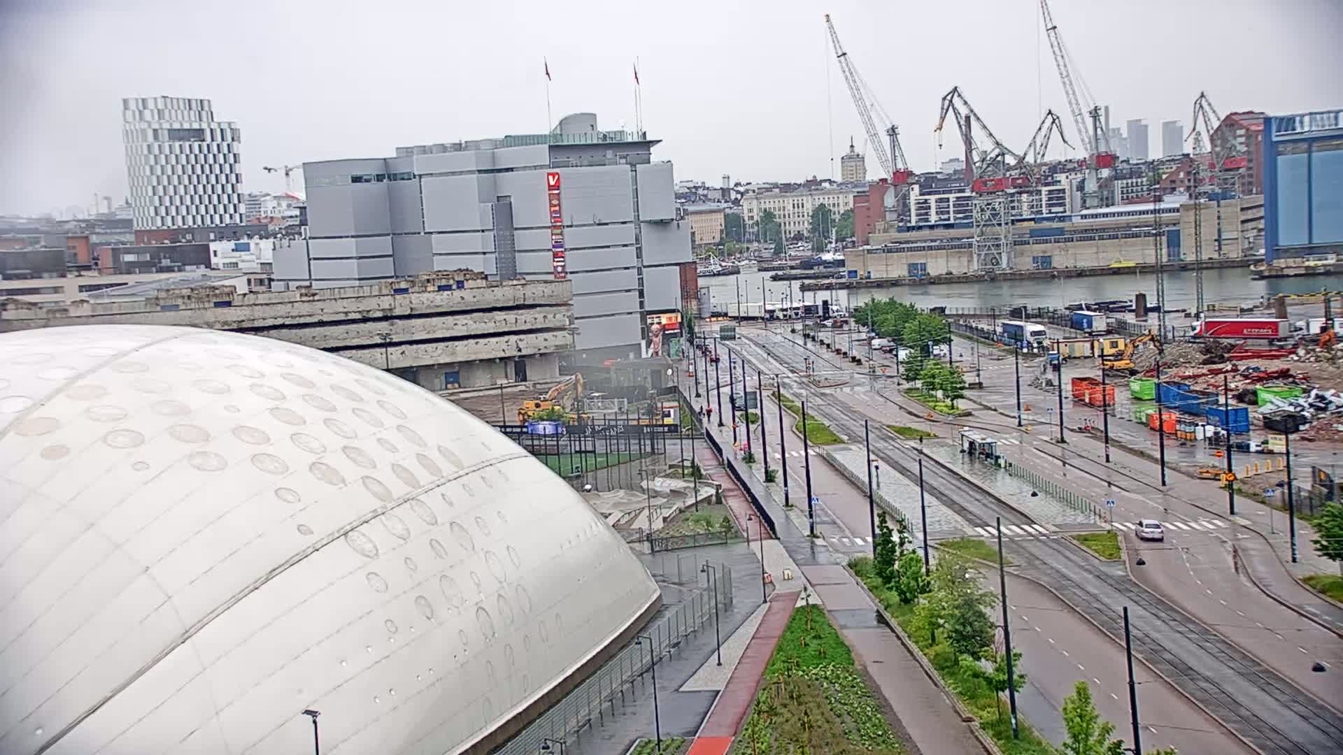 A large, domed building is visible in the foreground, with a cityscape including a harbor, construction sites, and tram lines in the background under overcast skies.