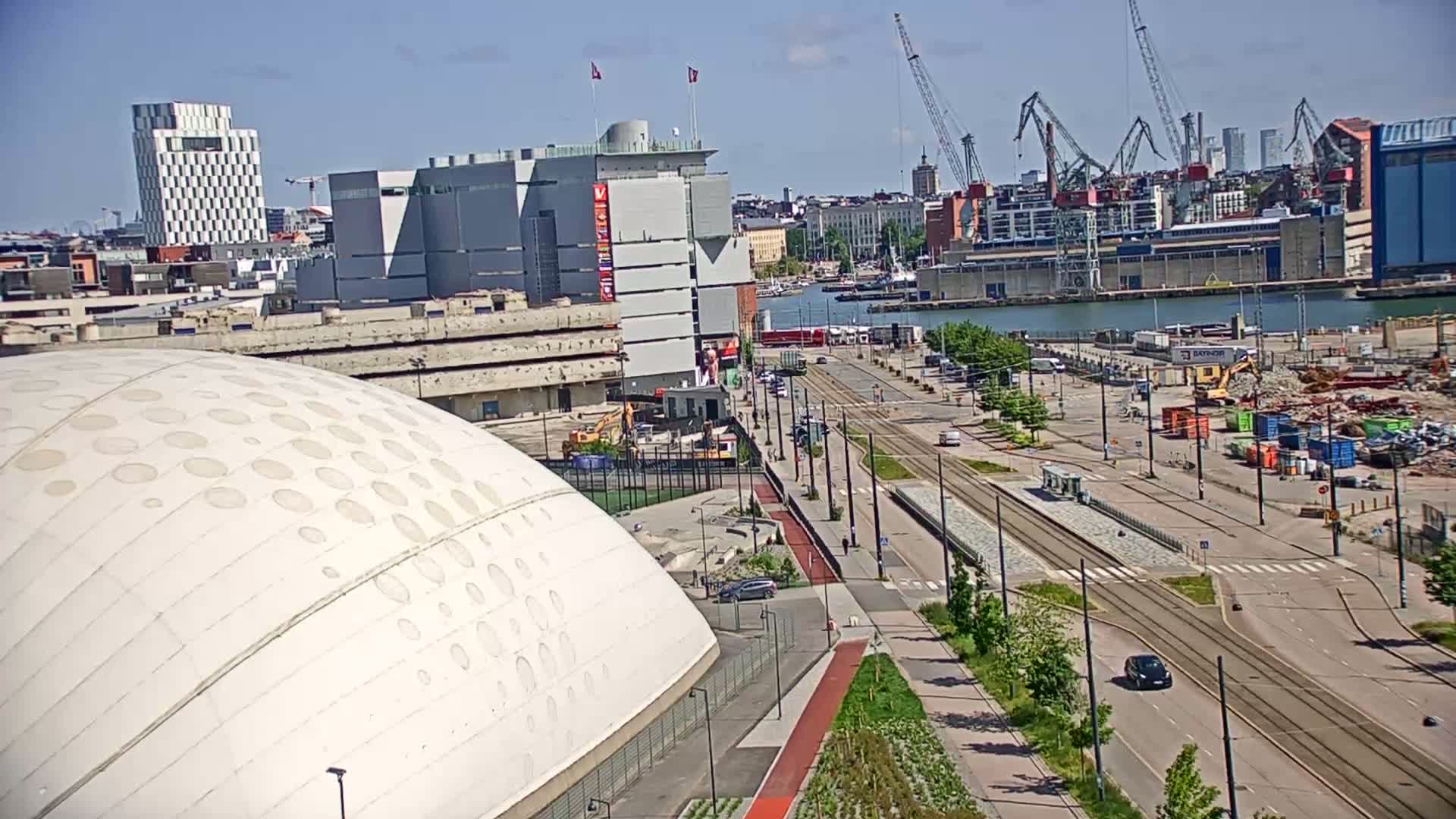 A large, domed building is situated near a harbor with construction and tram lines visible on a sunny day.