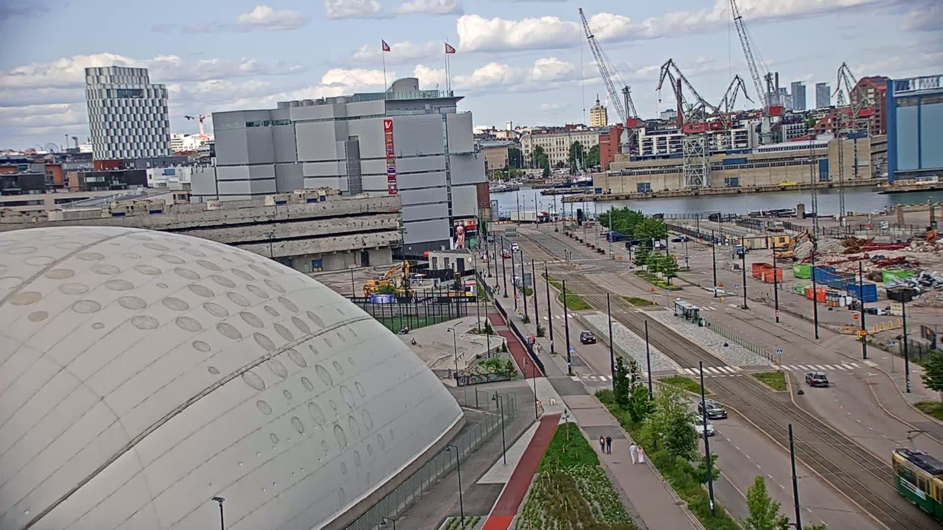 A large domed building sits beside a wide city street with tram tracks, under a partly cloudy sky, overlooking a harbor with cranes and ongoing construction.