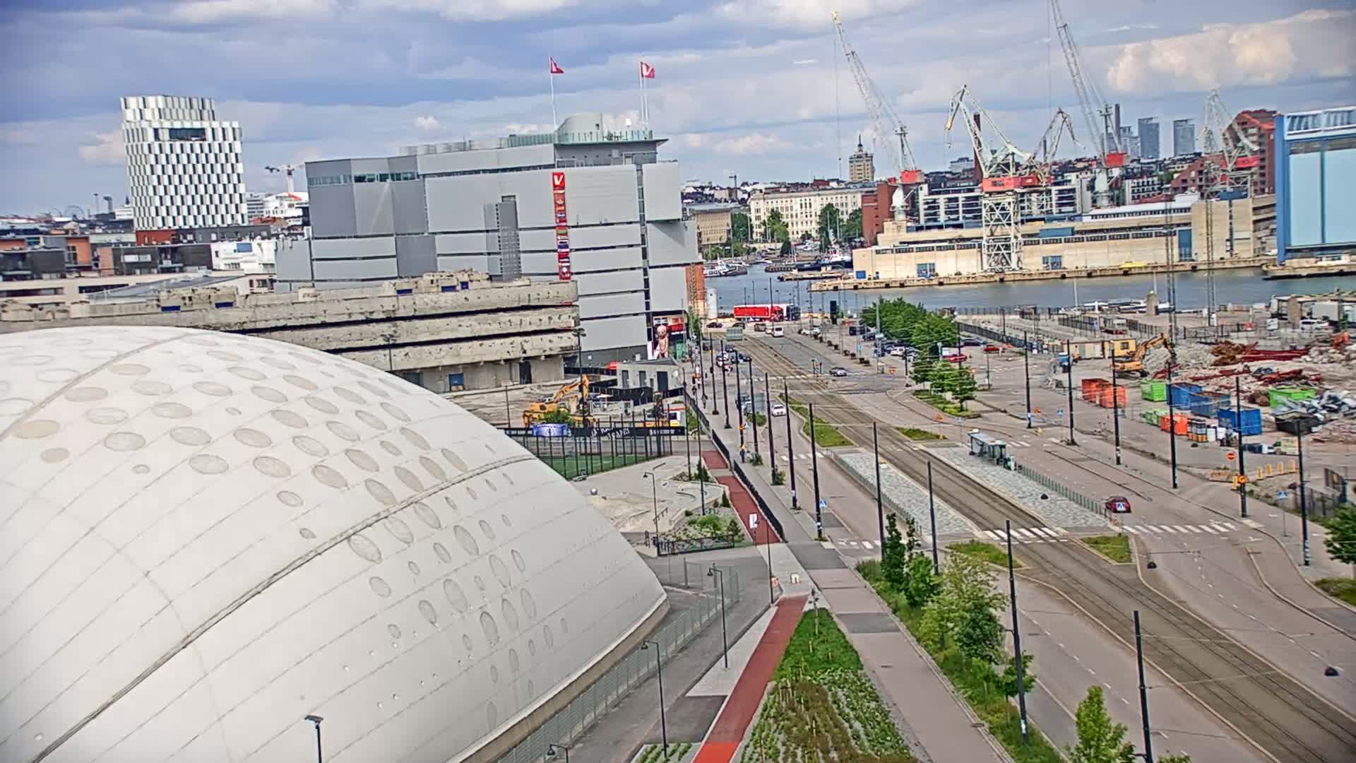 A large domed building sits beside a city street with tram tracks under partly cloudy skies, overlooking a harbor with cranes and construction activity.