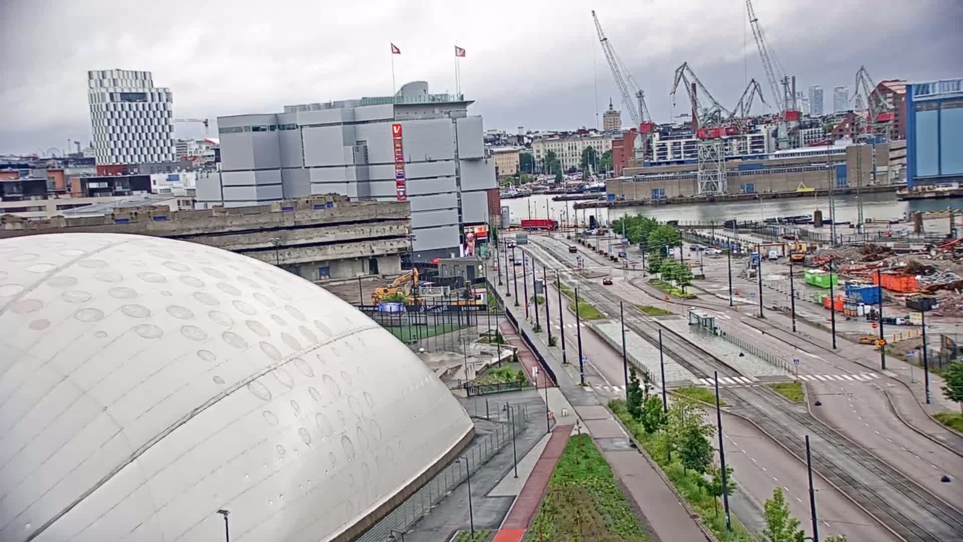 A large, domed building is situated near a city harbor on an overcast day, with construction and demolition visible nearby, and a wide road and tram tracks leading towards a waterfront with cranes and docked ships.