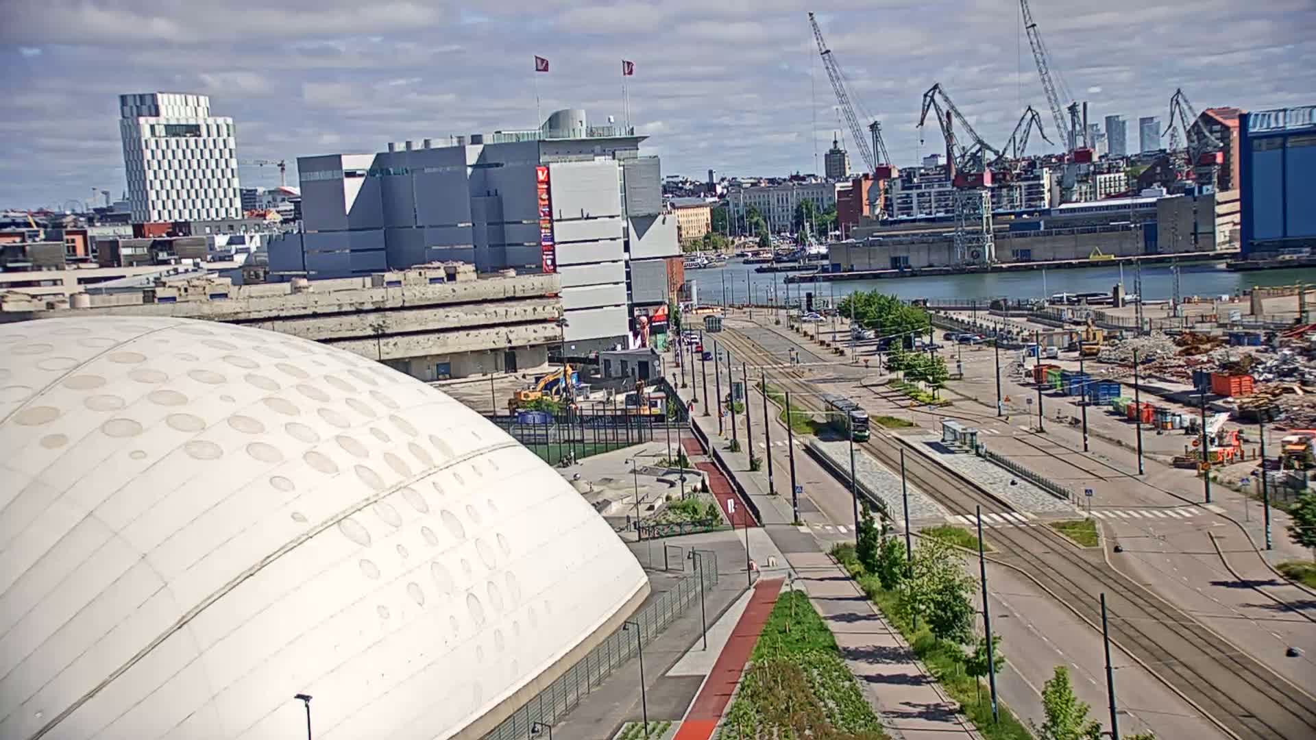 A large domed building sits beside a waterfront city scene under a partly cloudy sky, with tram tracks and construction visible in the foreground.