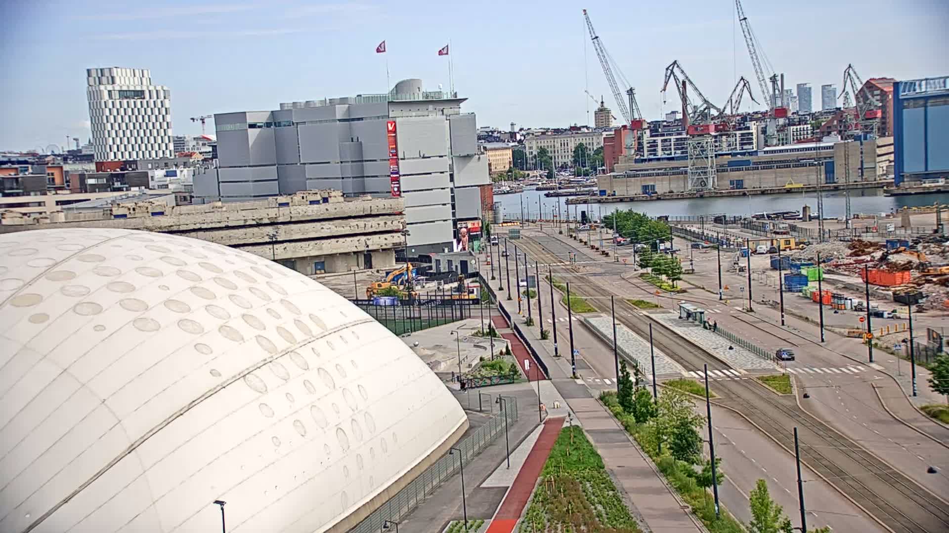 A large white domed building sits in the foreground of a city waterfront scene on a partly sunny day, with construction and cranes visible across a wide roadway and body of water.