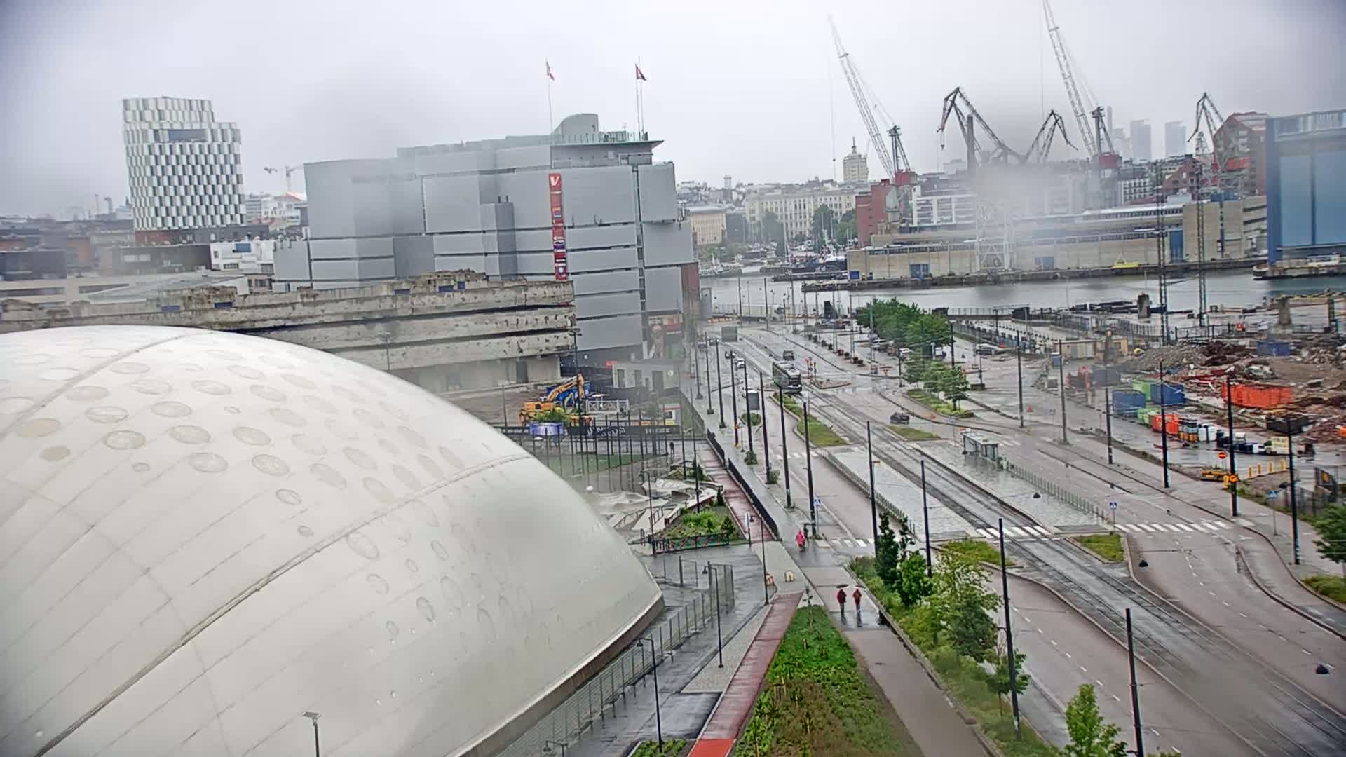 A large, domed building is seen in the foreground, with a city waterfront, cranes, and construction sites visible in the background under a gray, overcast sky.