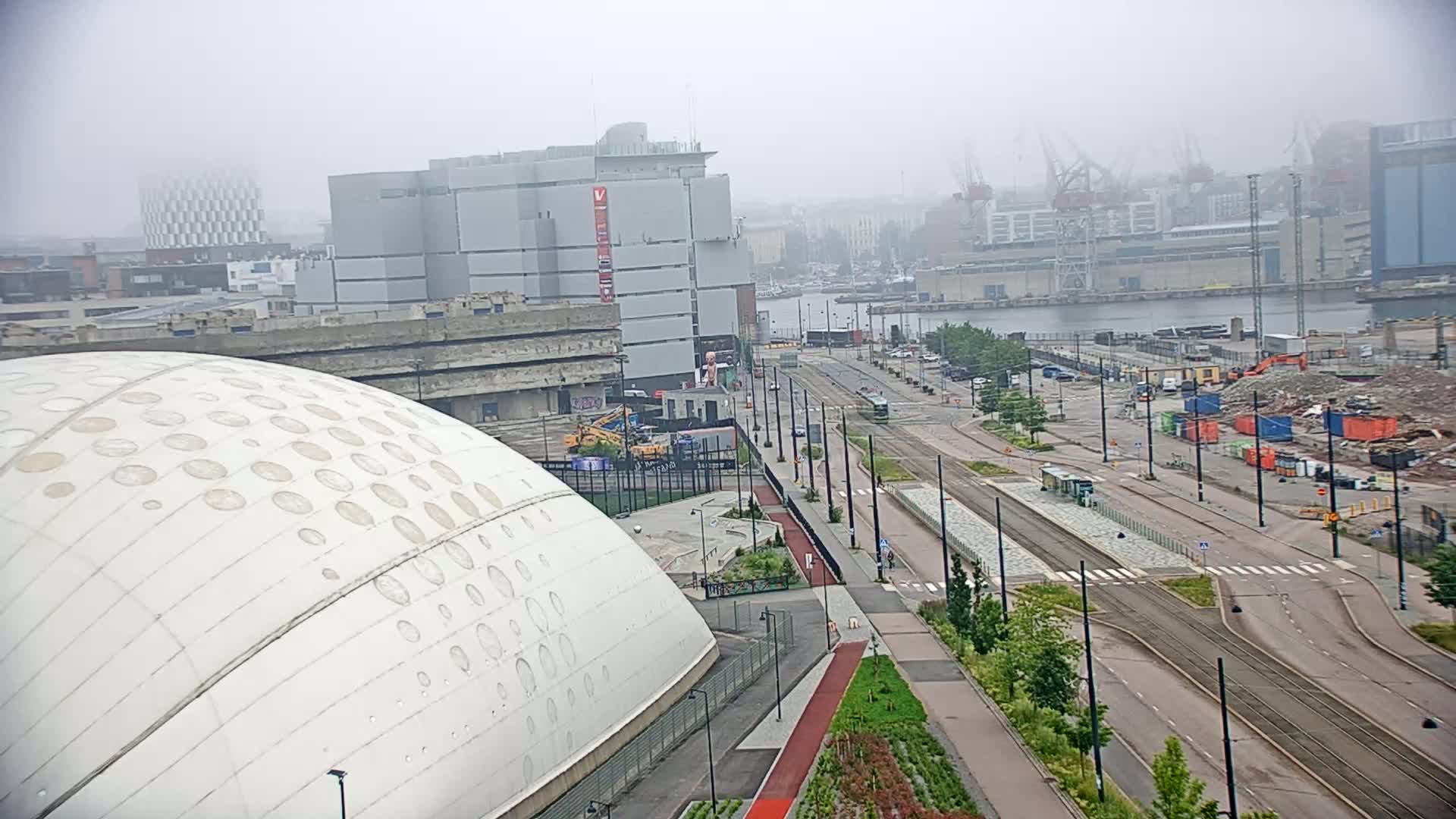 A foggy cityscape showing a large white dome-shaped building near a harbor with tram tracks and construction activity.