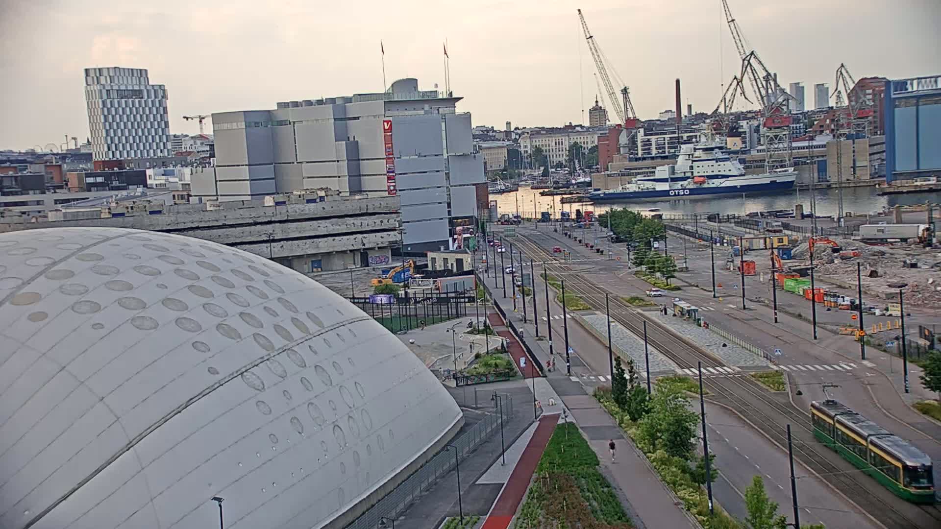 A large domed building sits next to a wide road with tram tracks and a harbor in the background under an overcast sky.