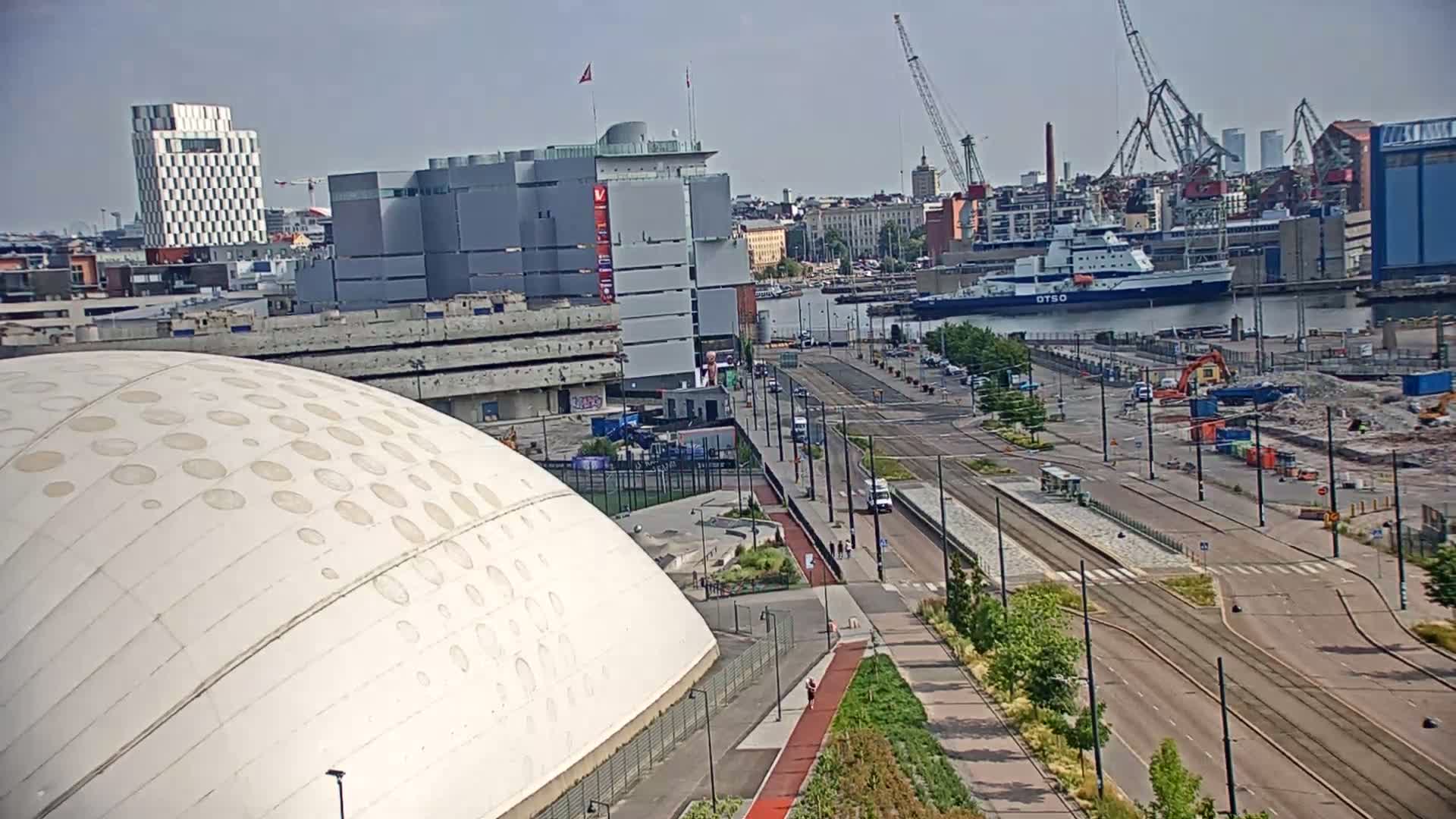 A large white domed building is situated near a harbor on a sunny day, with a road and tram lines running through a cityscape in the background.