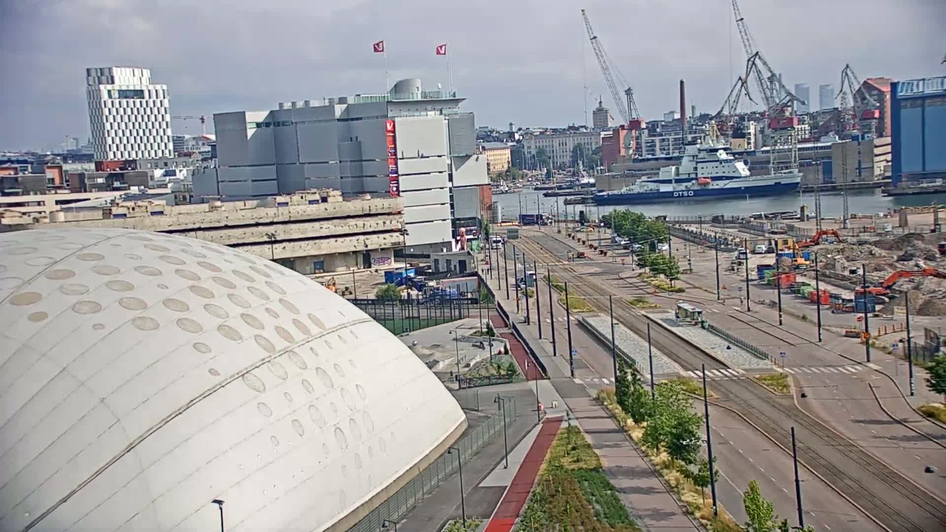 A large, domed building with circular patterns is seen next to a tram line and harbor with construction and ships under a mostly cloudy sky.