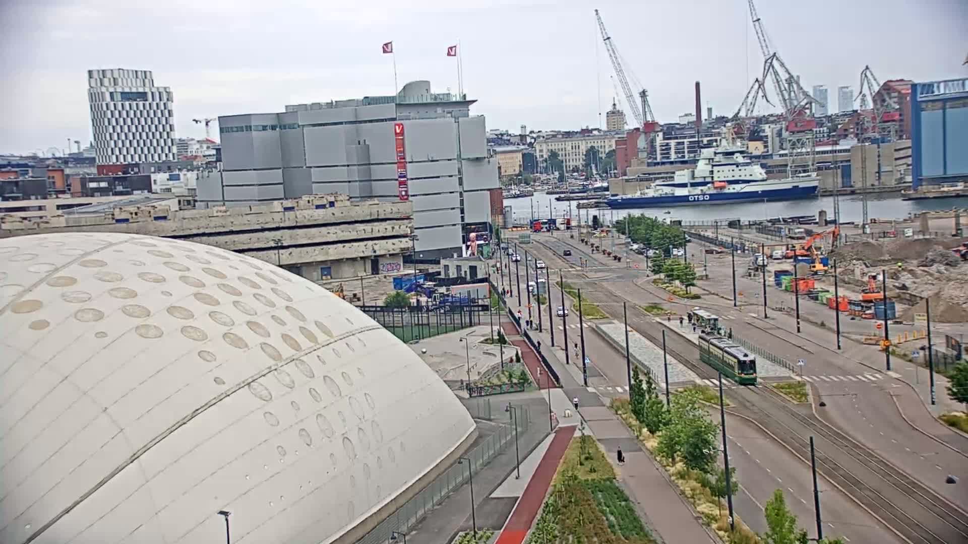 A large, domed building with circular patterns is visible in the foreground, next to a city street with a tram and various buildings, construction sites, and a body of water with ships in the background under an overcast sky.