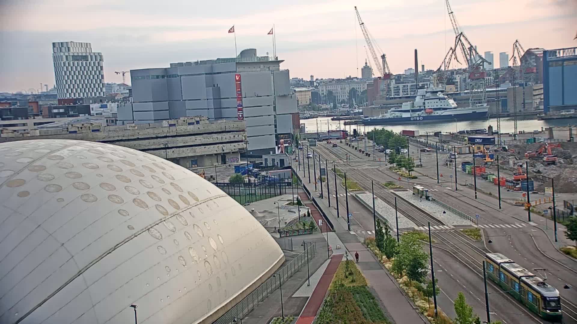 A large, domed building with circular patterns is visible in the foreground, next to a city street with a tram and various buildings, construction sites, and a body of water with ships in the background under an overcast sky.