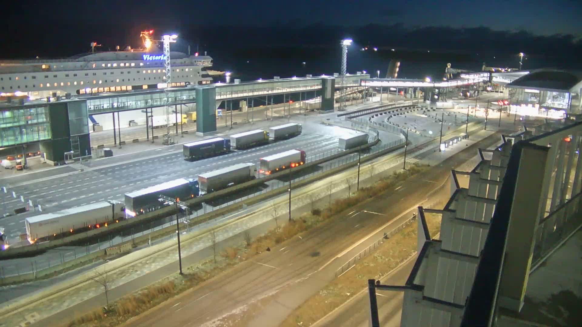 A large, domed building with circular patterns is visible in the foreground, next to a city street with a tram and various buildings, construction sites, and a body of water with ships in the background under an overcast sky.