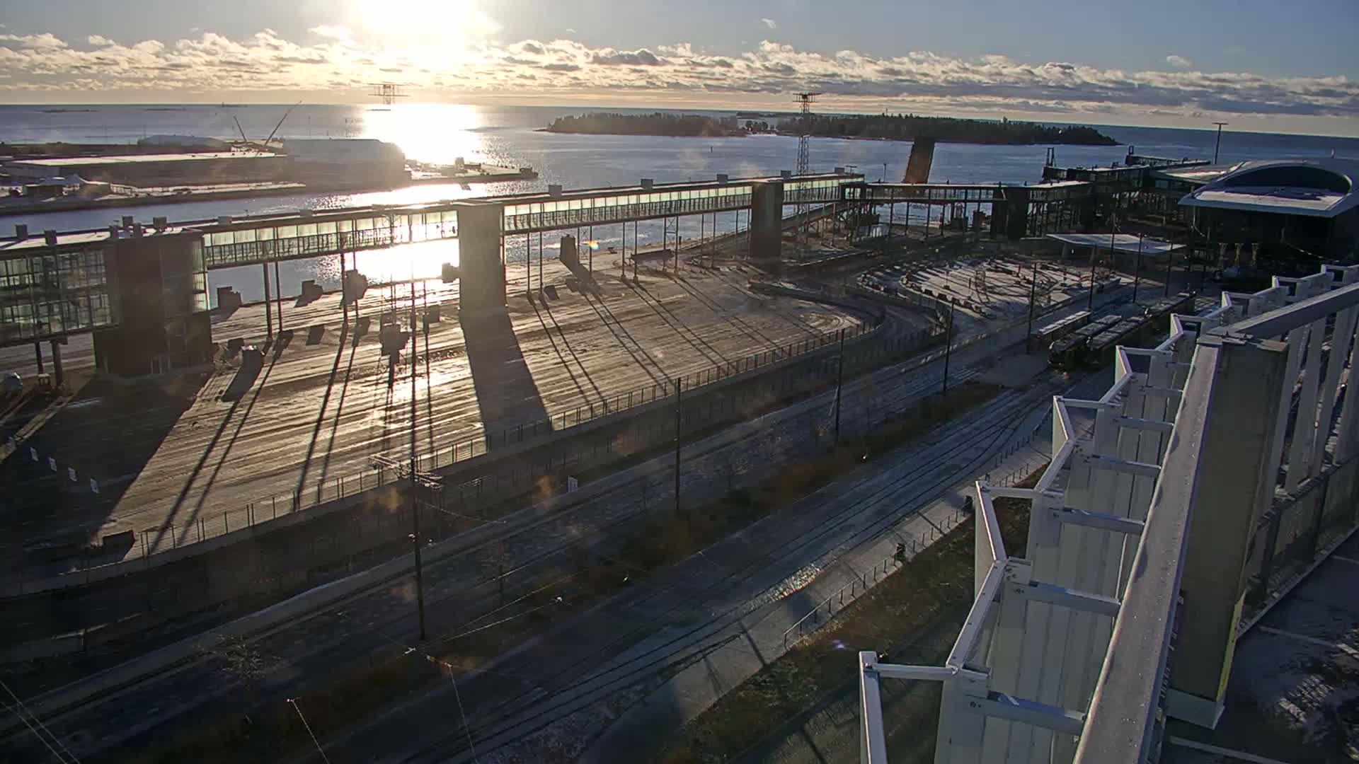 A modern port or ferry terminal is seen under bright sunlight with scattered clouds, showcasing elevated walkways connecting buildings, vast paved areas with train tracks, and distant islands, all casting prominent long shadows.