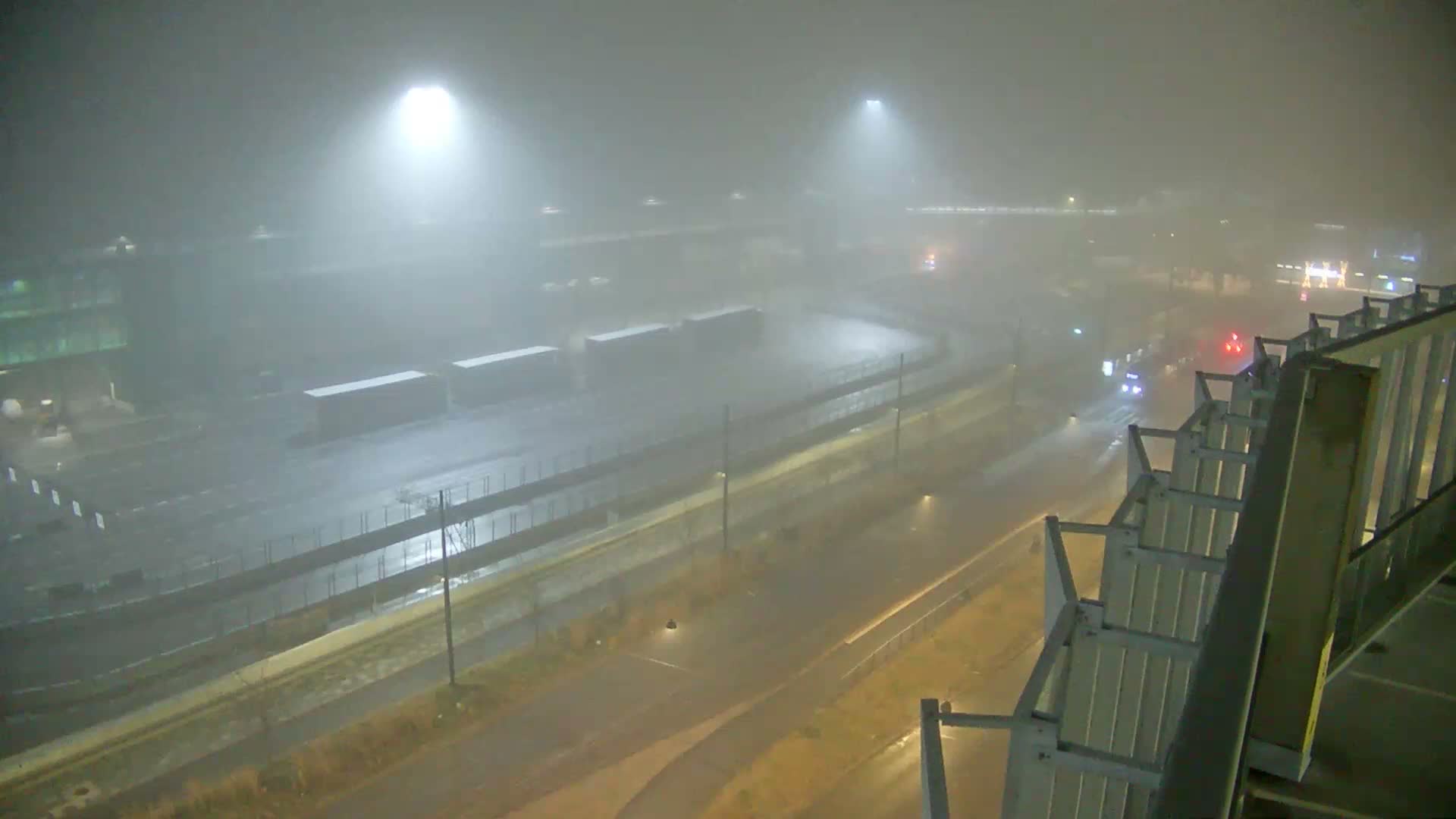 An elevated view captures a sprawling, wet industrial or transportation hub at night, heavily shrouded in fog, with bright floodlights illuminating containers and a road where distant vehicles are visible, framed by a building's railing on the right.