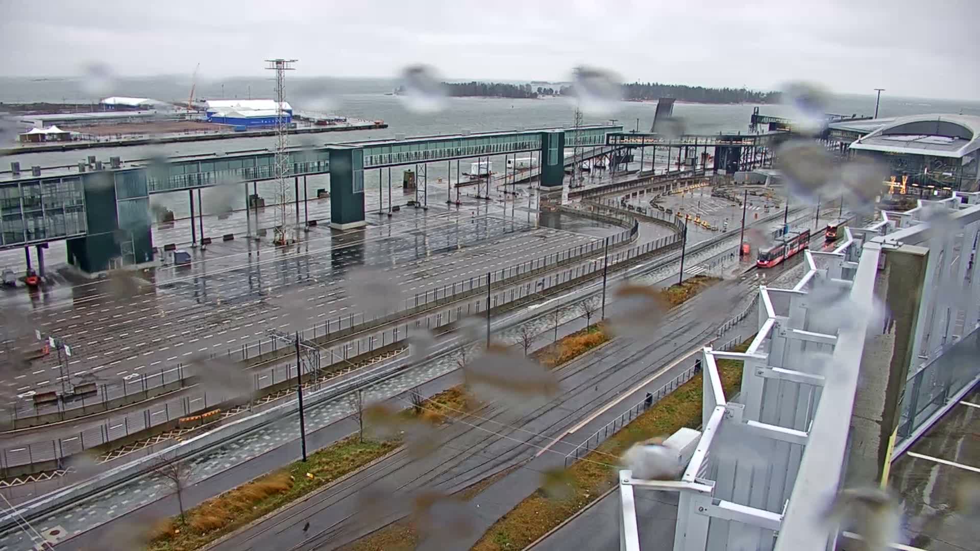 A high-angle view captures a rainy, overcast day at a modern port or ferry terminal, featuring wet asphalt, tram tracks with a red tram, an elevated walkway, and the choppy sea in the background, all viewed through a rain-splashed lens.