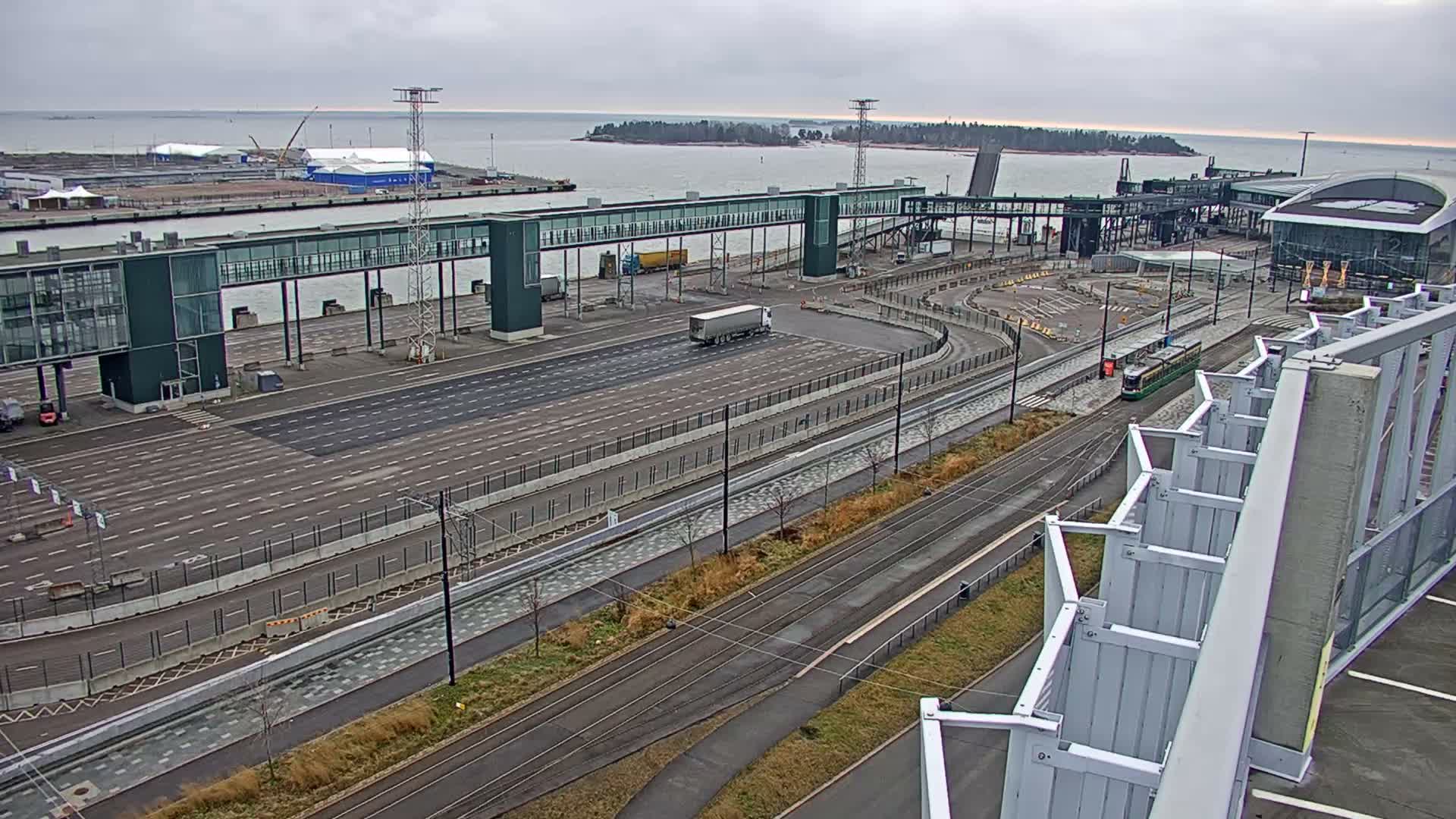 An overcast and grey day reveals a busy modern port or ferry terminal with wide multi-lane roads, a white semi-truck, a green tram on tracks, several large buildings, and distant islands across the water.
