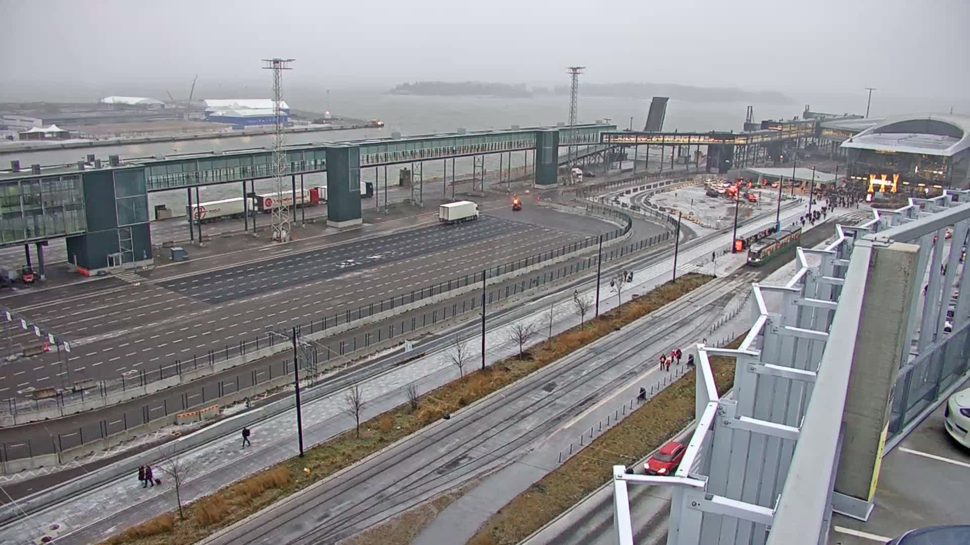 An overcast and grey day reveals a busy modern port or ferry terminal with wide multi-lane roads, a white semi-truck, a green tram on tracks, several large buildings, and distant islands across the water.