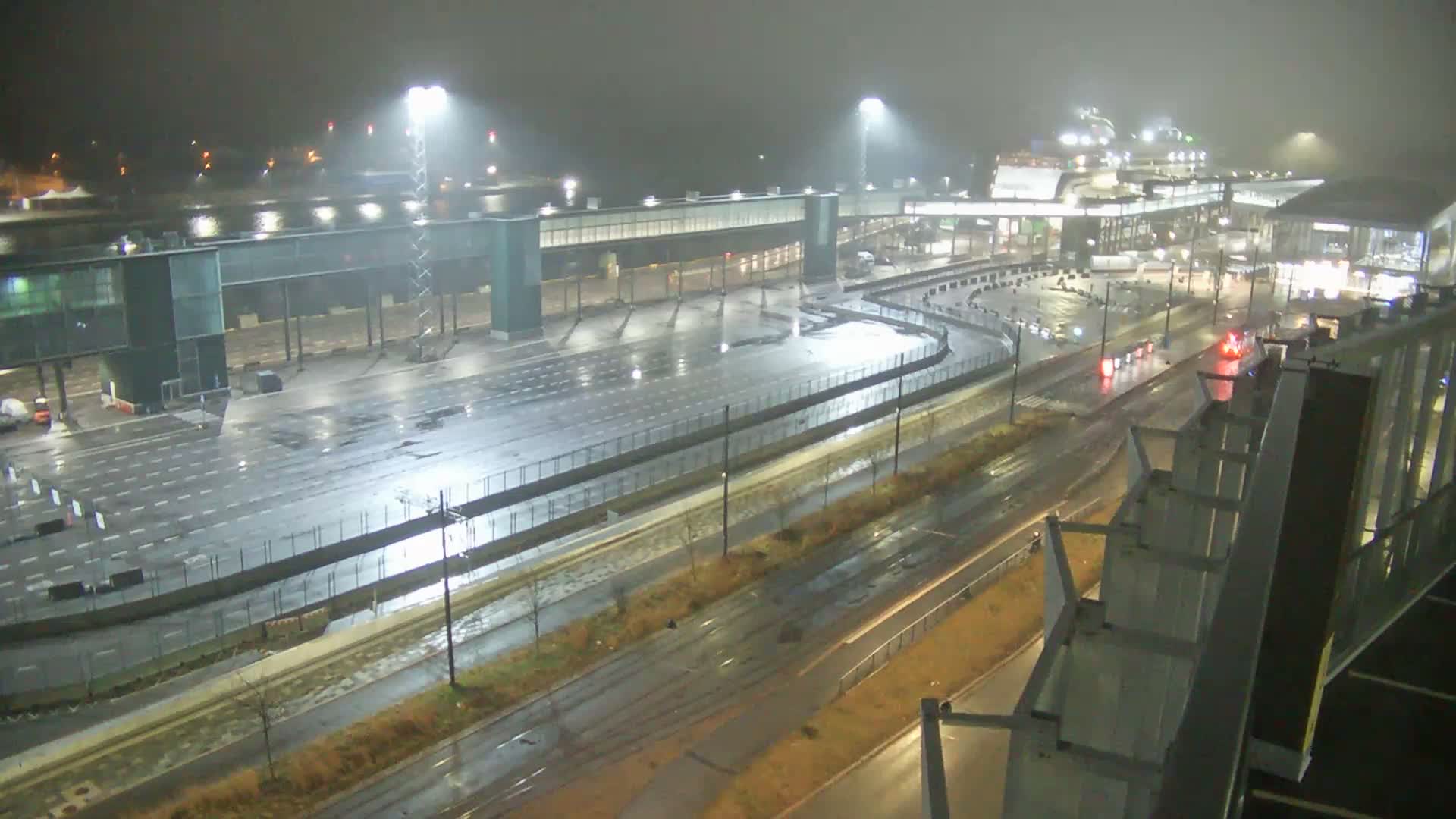 An overcast and grey day reveals a busy modern port or ferry terminal with wide multi-lane roads, a white semi-truck, a green tram on tracks, several large buildings, and distant islands across the water.