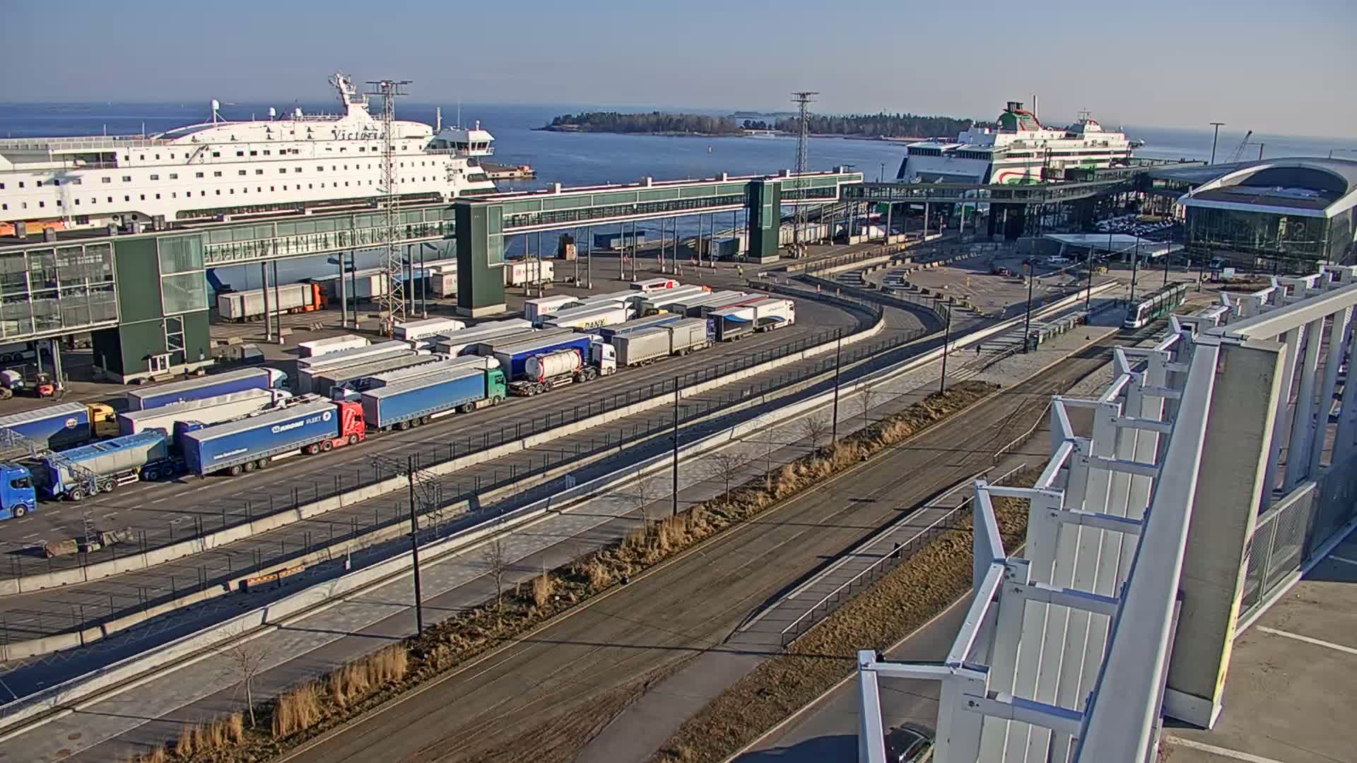 An overcast and grey day reveals a busy modern port or ferry terminal with wide multi-lane roads, a white semi-truck, a green tram on tracks, several large buildings, and distant islands across the water.