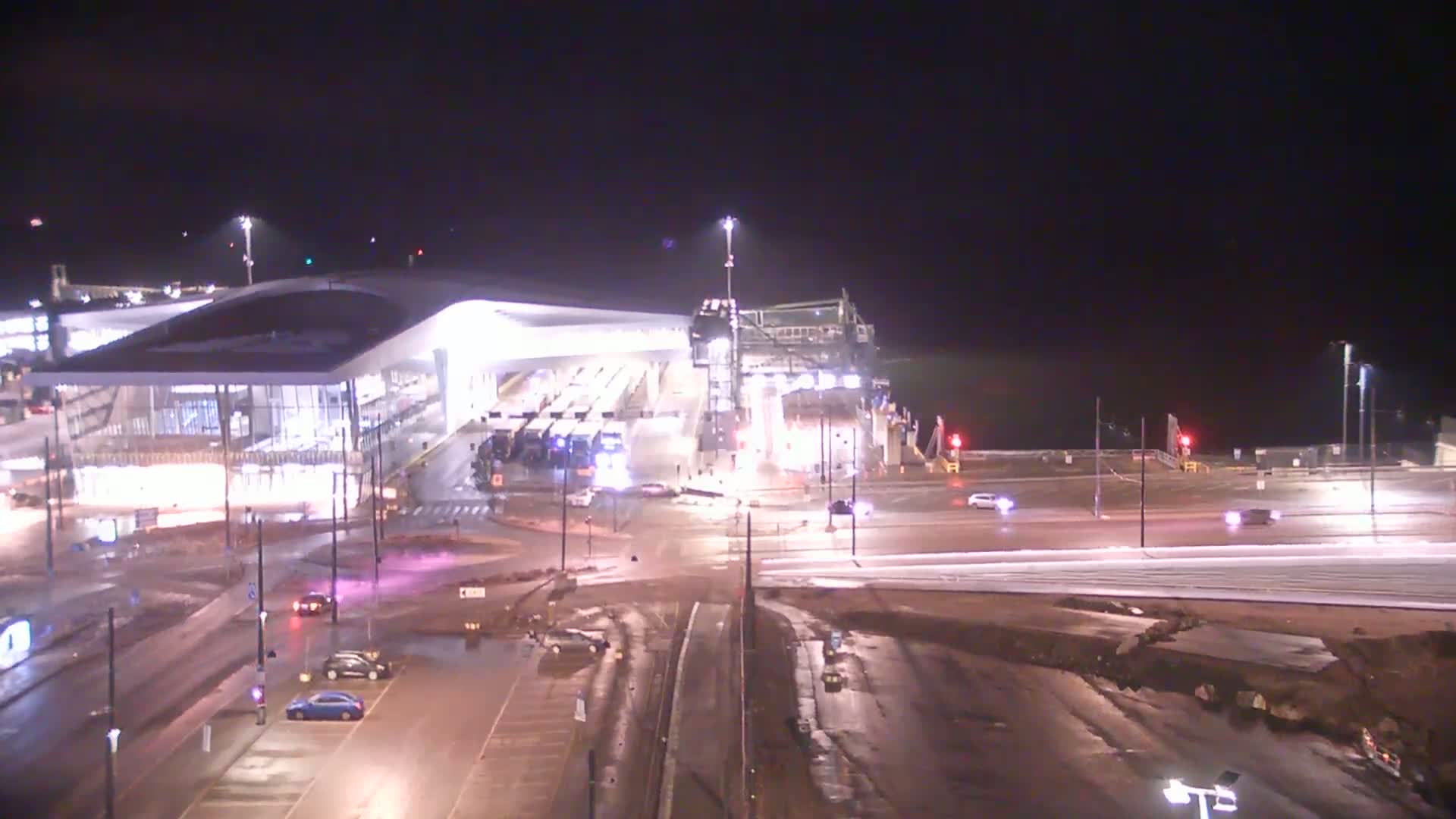 A brightly lit ferry terminal at night, with several vehicles present and wet roads.