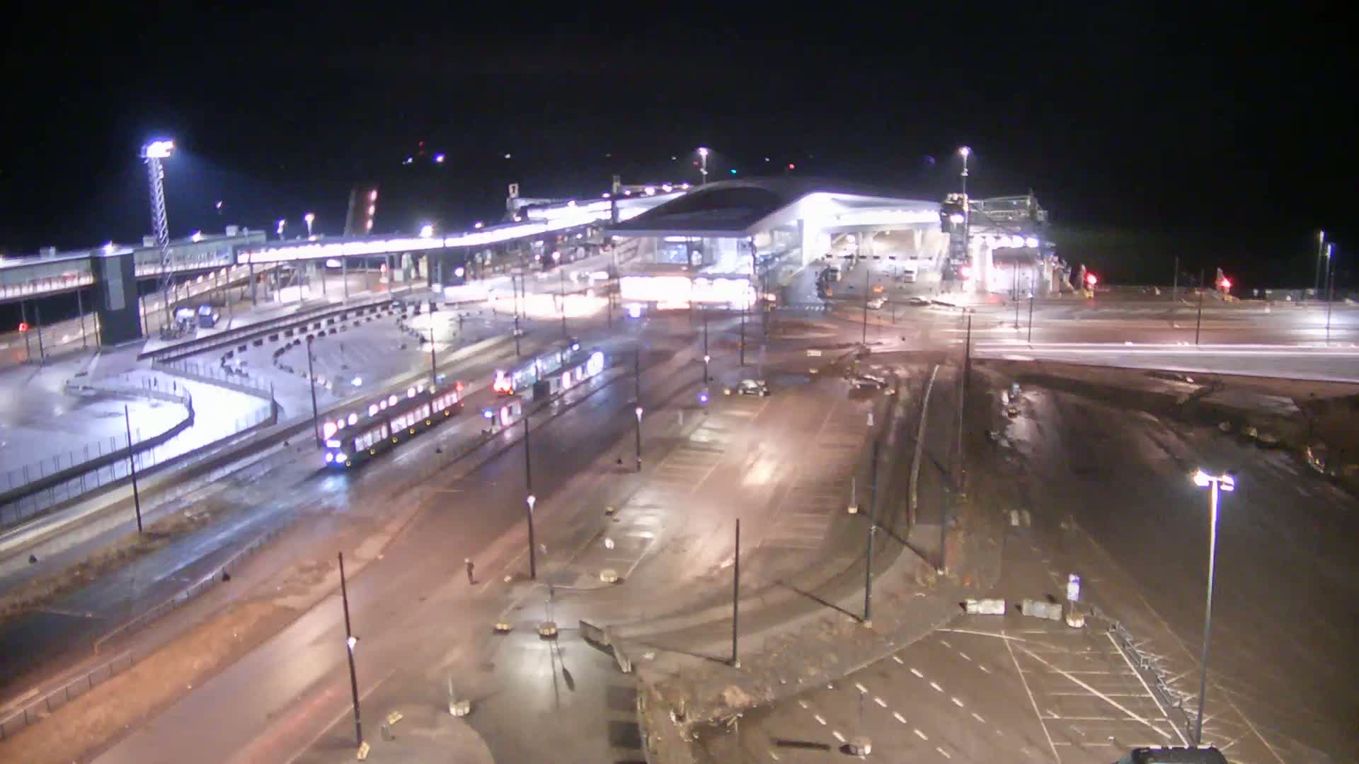 A nighttime, wet scene of a transportation hub with a large modern building, roadways, and a tram, with some construction or maintenance visible.
