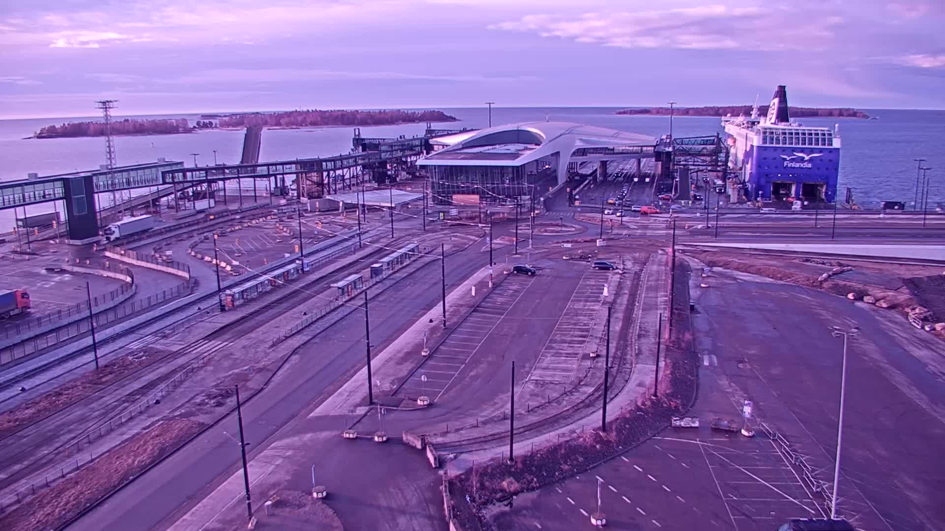 A ferry is docked at a terminal with a passenger walkway, parking lots, and tram tracks, under a purplish twilight sky.