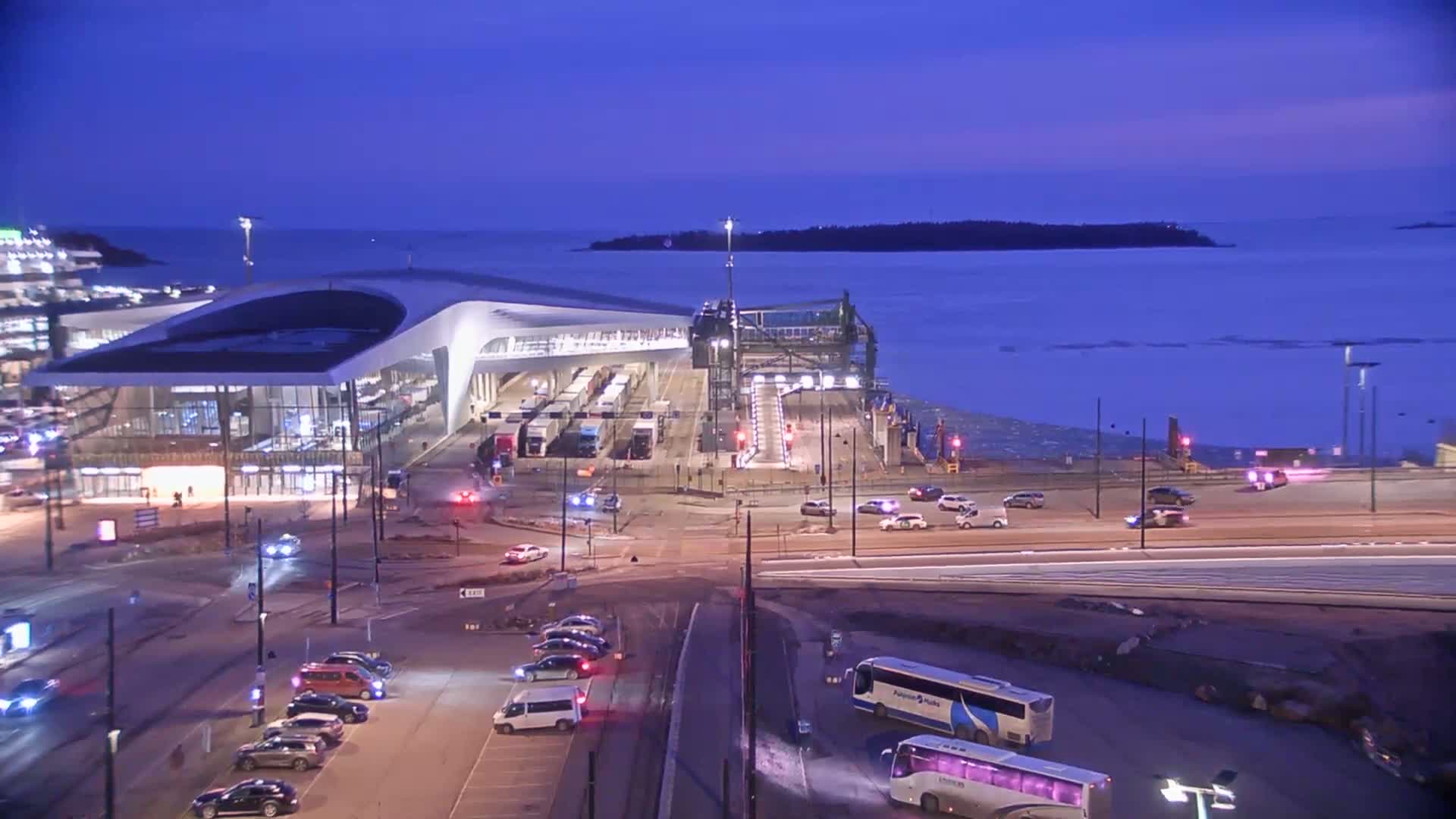 A modern ferry terminal with a curved roof is illuminated at dusk, overlooking a calm body of water partially covered in ice, with vehicles on roads surrounding the terminal.