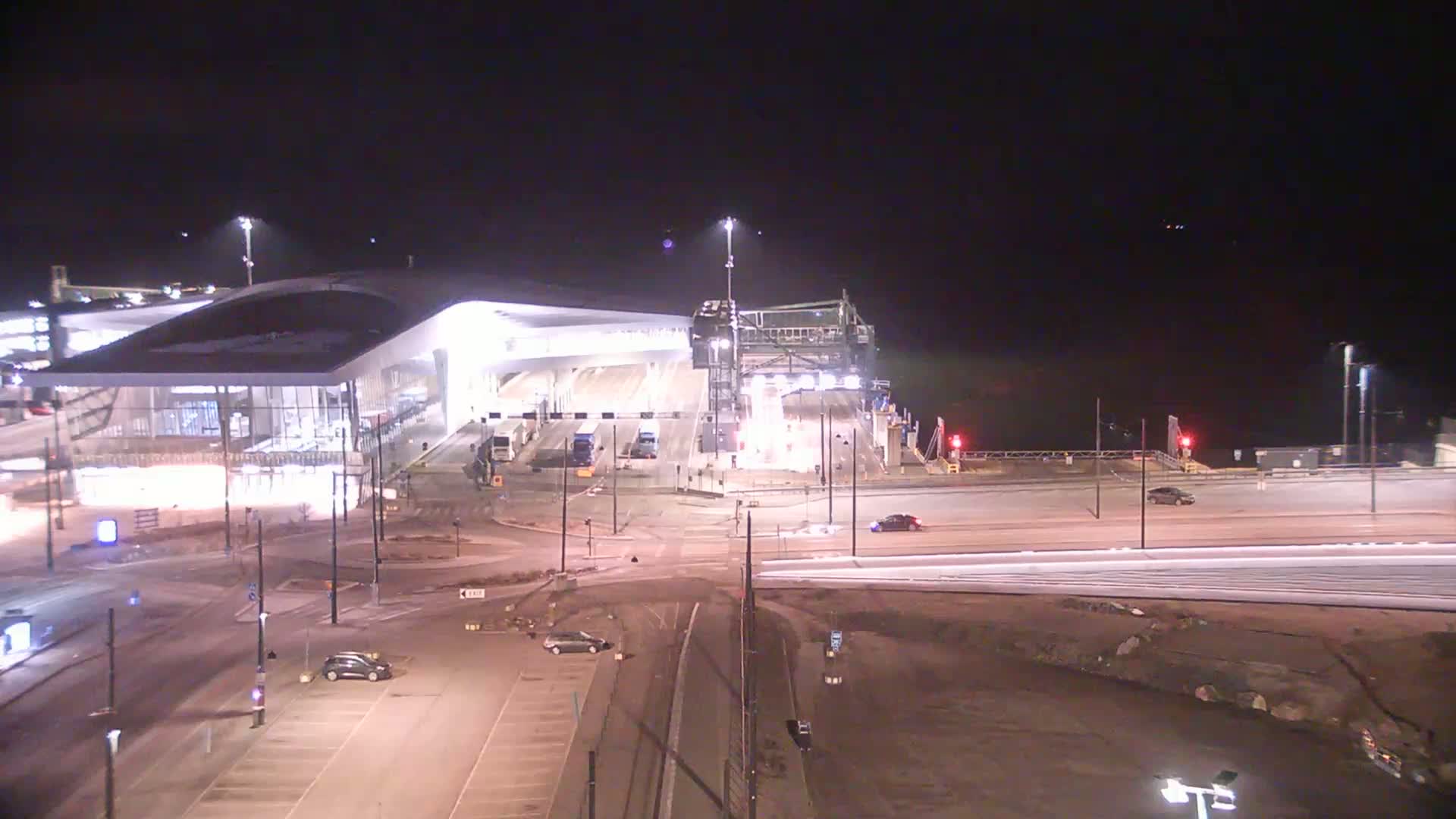 A brightly lit ferry terminal and surrounding area at night, with several cars visible in the parking lot and on nearby roads.