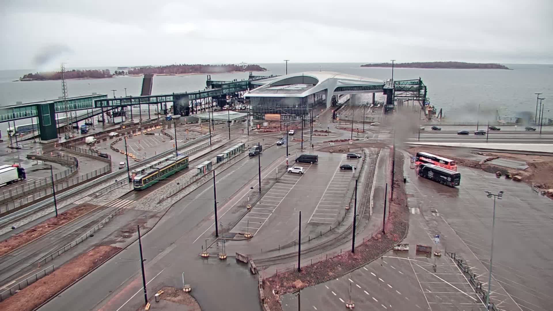A ferry terminal with a tram and several vehicles is seen on a rainy day, with a body of water and islands in the background.