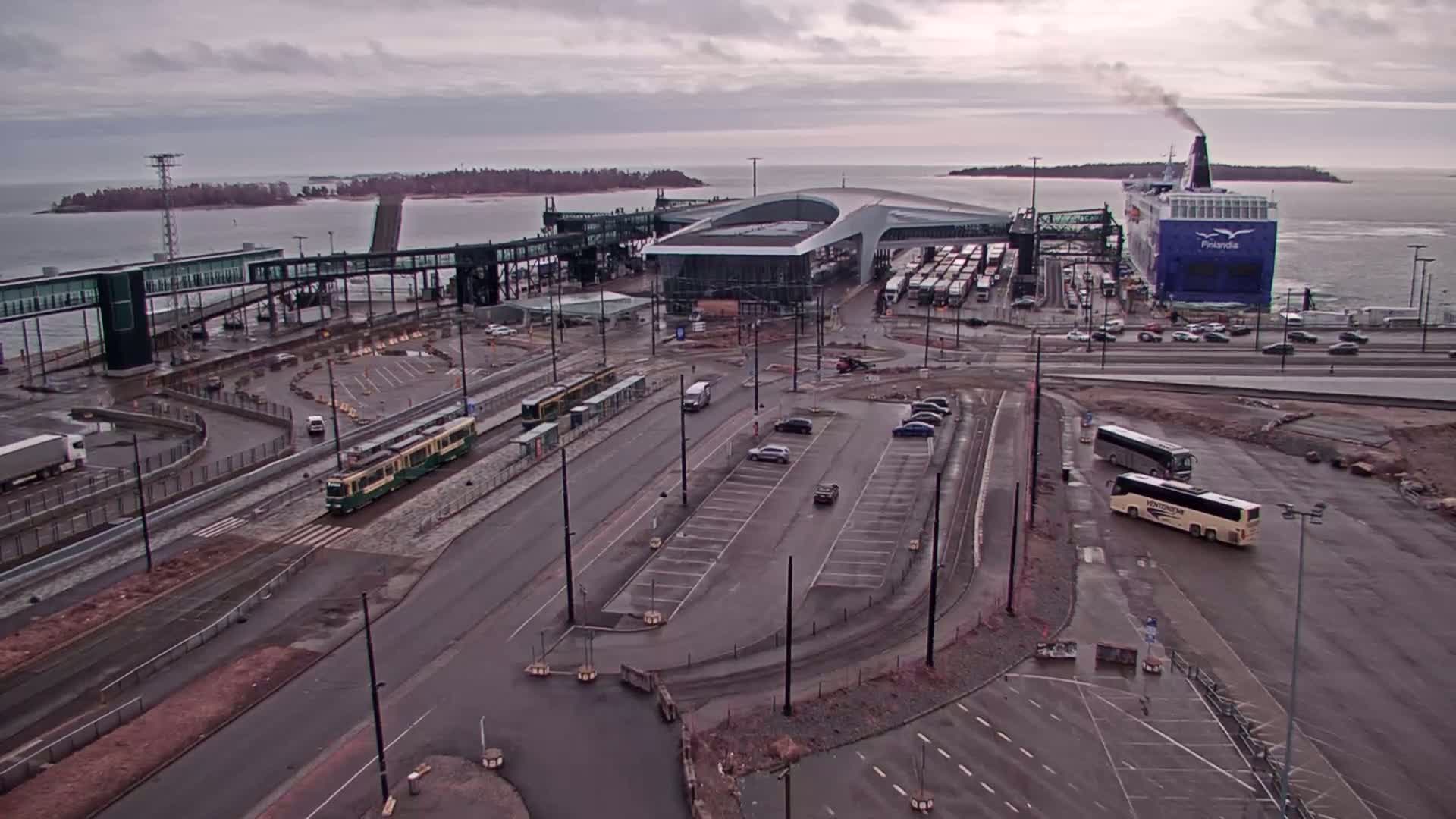 An aerial view of a ferry terminal on an overcast day shows a ferry docked at the terminal, trams and buses parked nearby, and cars in a parking lot.