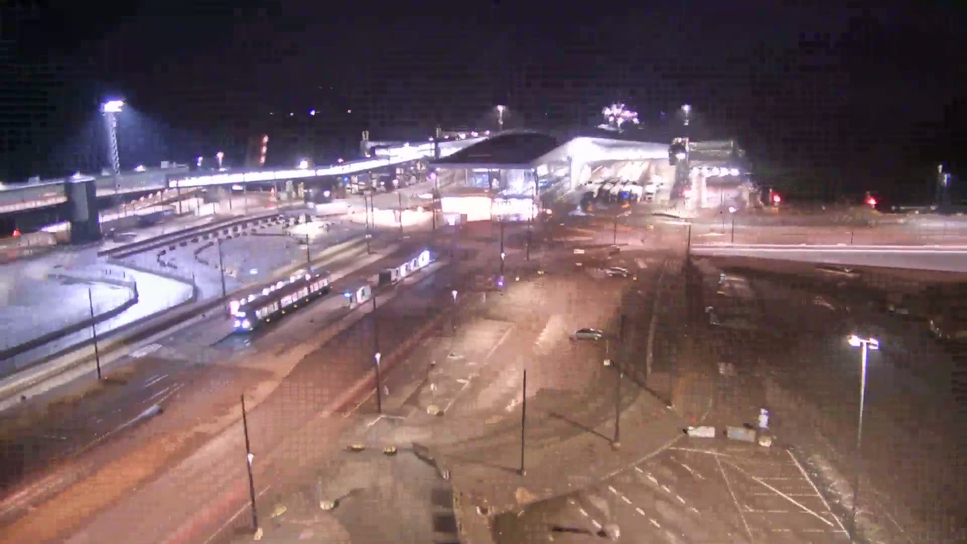 A nighttime, overhead view shows a transportation hub with a train and several vehicles in a large, mostly empty parking lot.