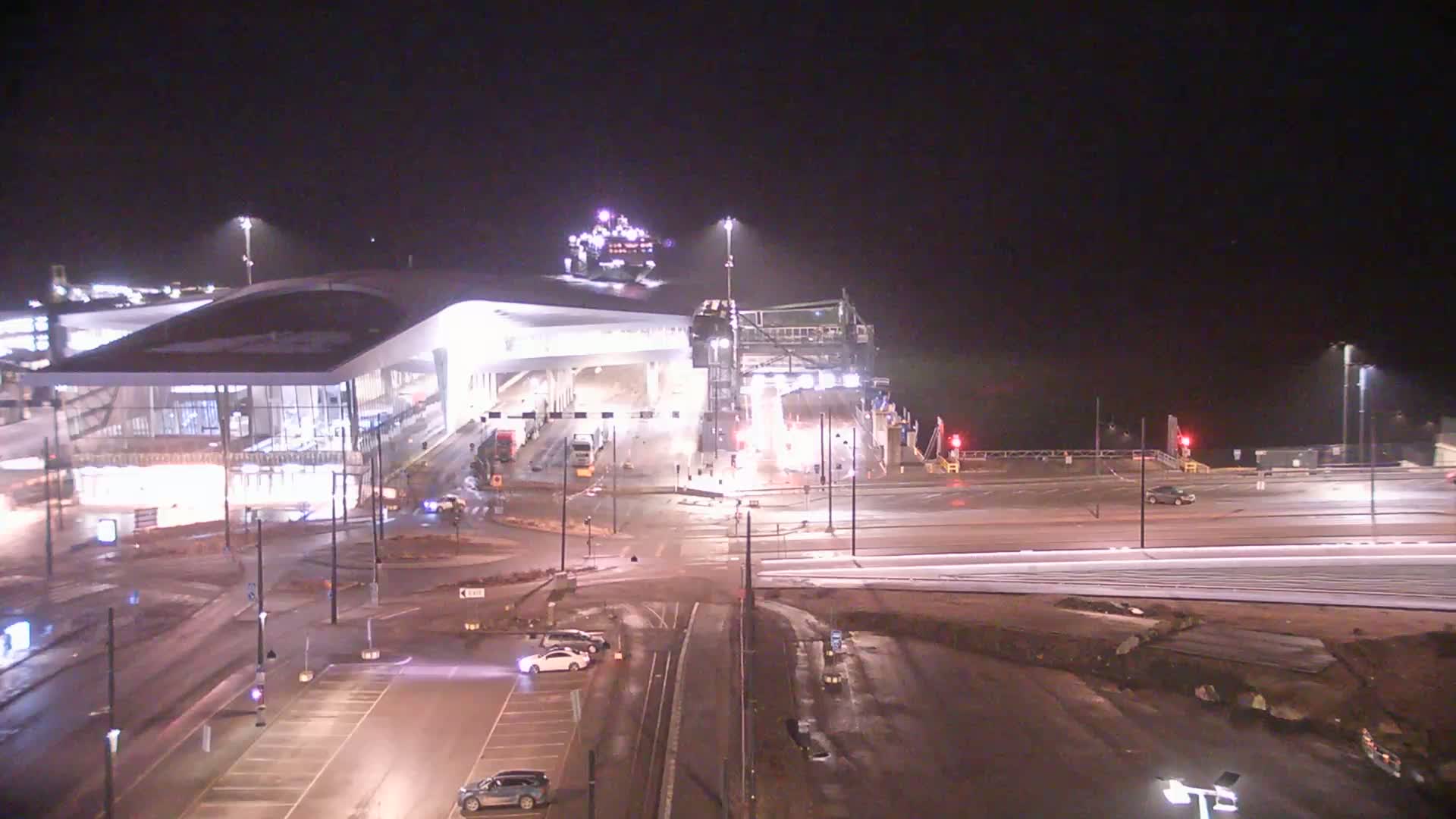 A modern ferry terminal at night, illuminated under artificial lights, with a ferry docked and several vehicles in the surrounding area.