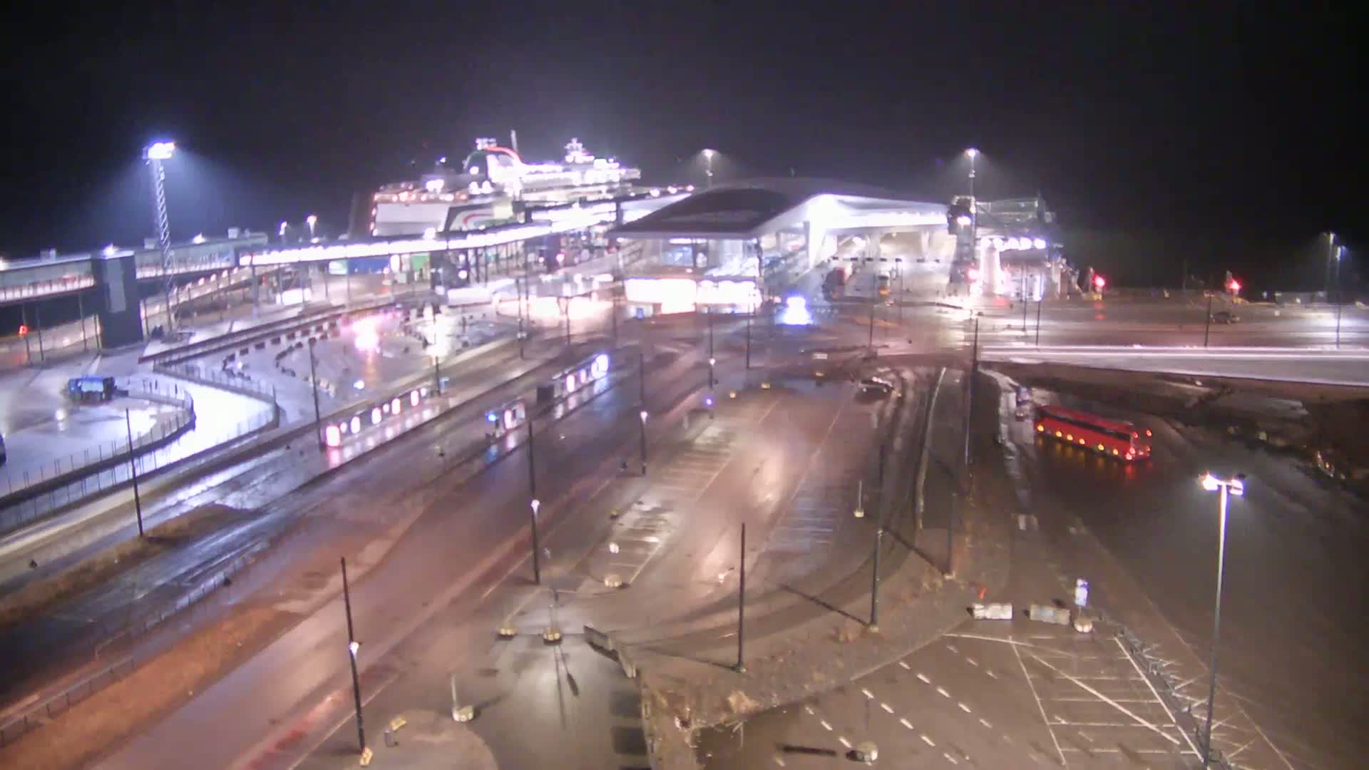 A ferry is docked at a terminal at night in wet conditions; several vehicles are visible on the surrounding roads and parking lots.