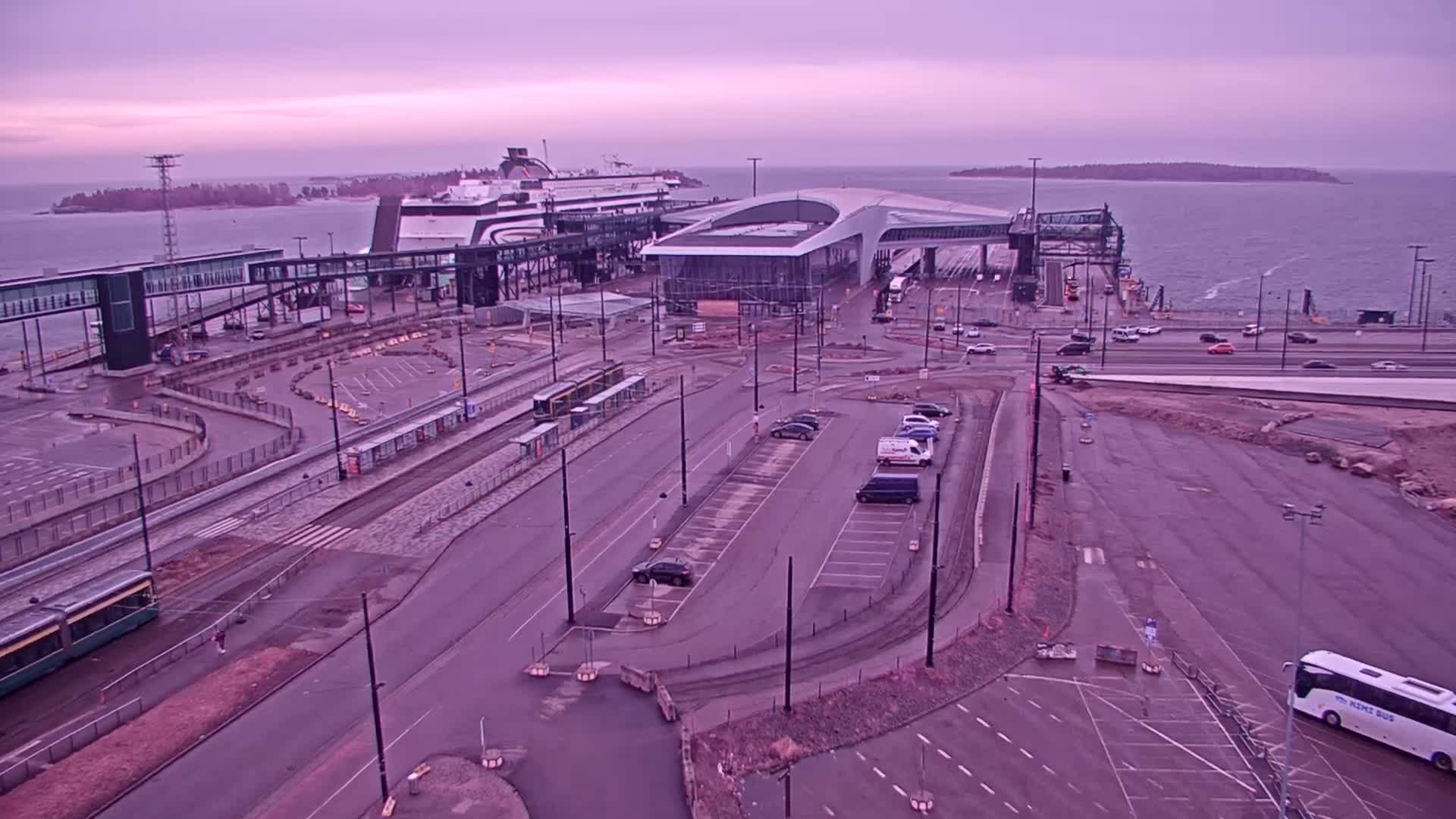 An aerial view of a ferry terminal with a large ferry docked, trams, cars in parking lots, and a bus under an overcast sky.