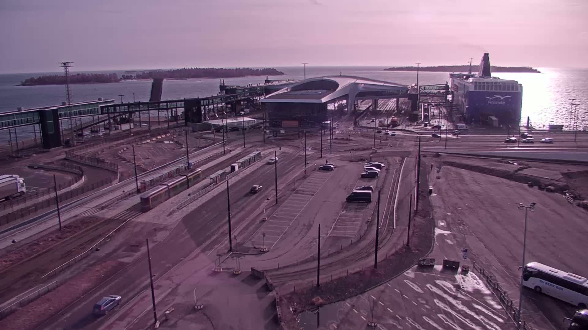 A ferry is docked at a terminal with a tram line and parking lot visible under a bright, somewhat hazy sky.