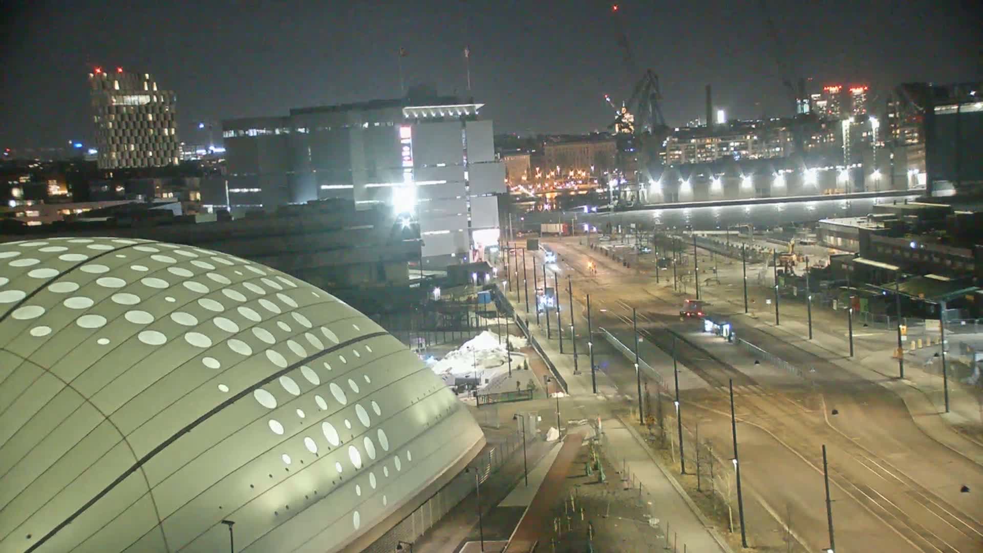 A nighttime, high-angle view of a city street with tram tracks, buildings, and a domed structure patterned with white circles.