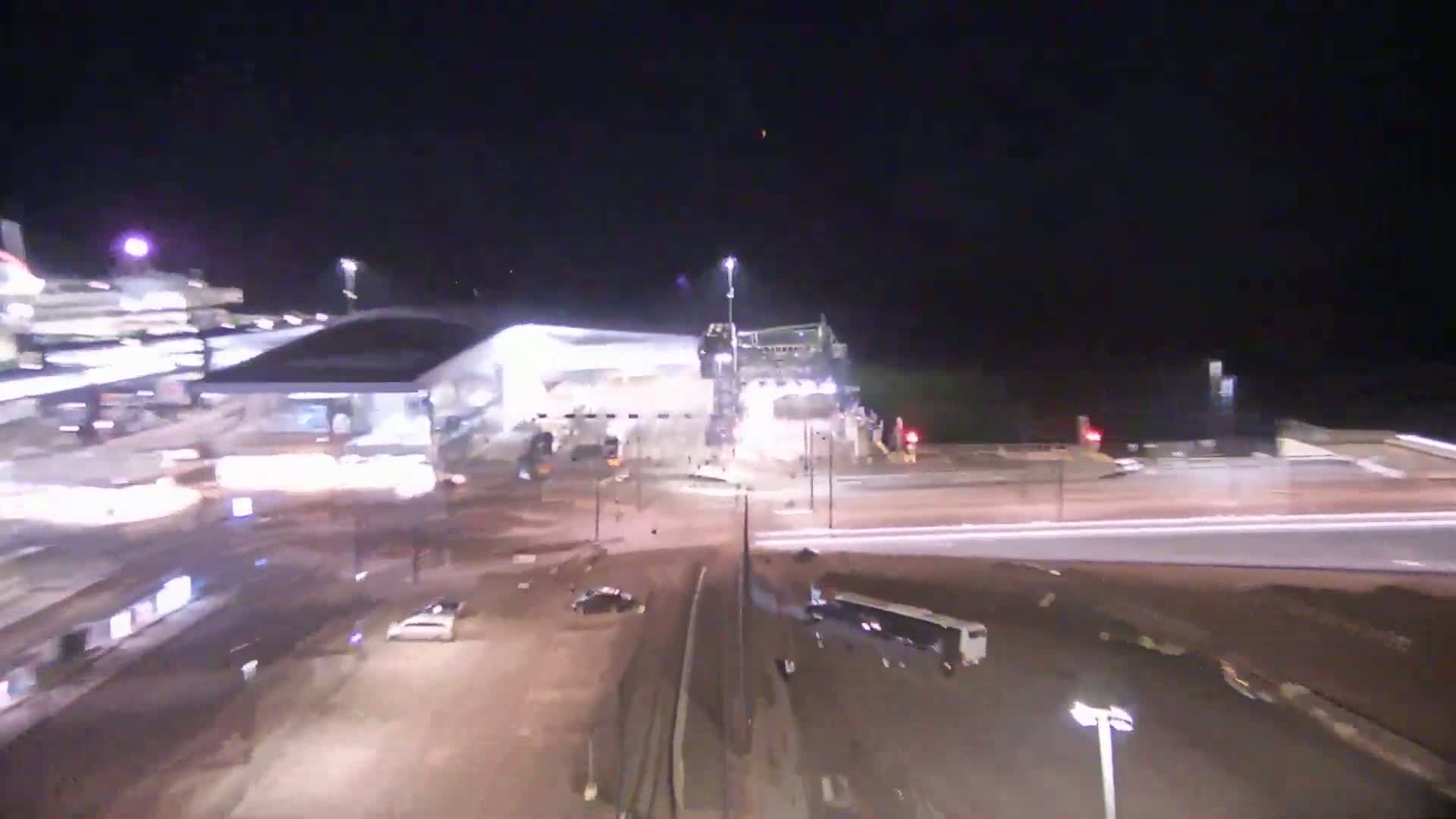 A nighttime aerial view shows a large building, vehicles in a parking lot and on roadways, and some construction activity under clear skies.