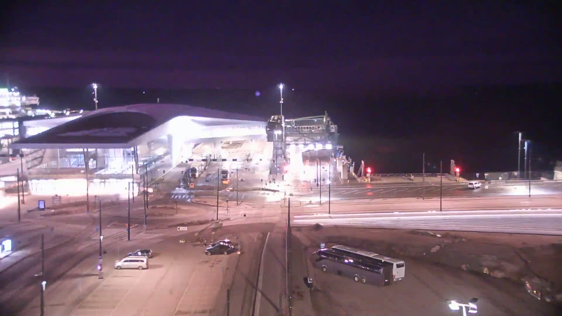 A brightly lit ferry terminal at night, with a few cars and a bus in the parking lot, under a dark, clear sky.