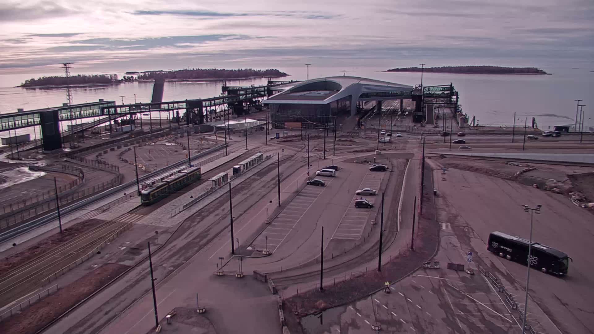 A ferry terminal with a tram, several cars in a parking lot, and a bus are visible on an overcast day, overlooking a calm body of water and islands in the distance.
