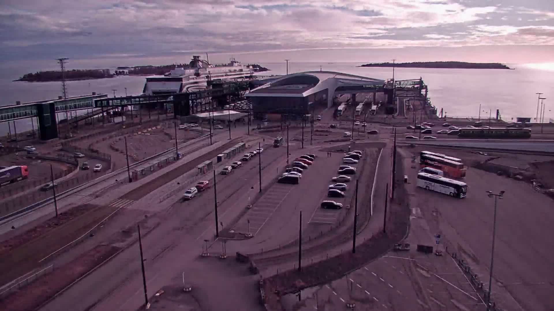 An aerial view of a ferry terminal with a large ferry docked, numerous cars parked in a lot, and several buses, under a cloudy sunset sky.
