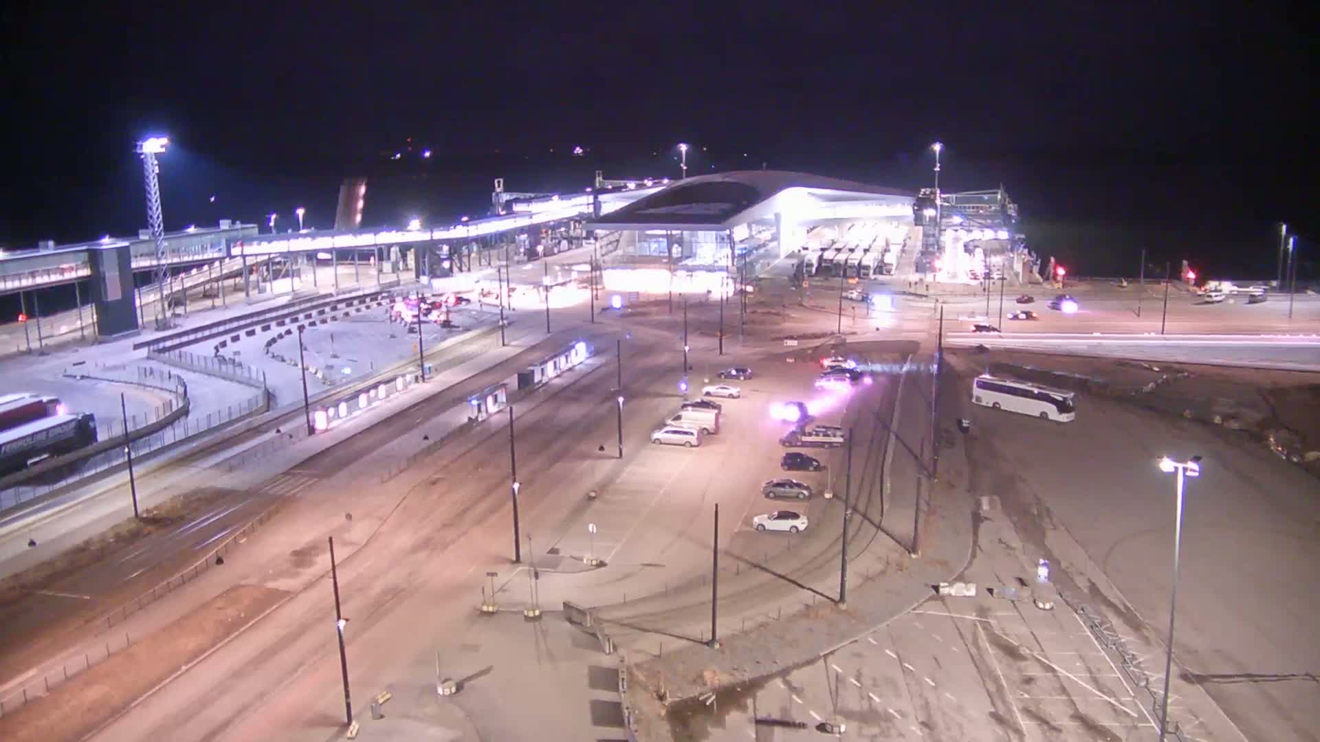 A nighttime aerial view shows a large, modern terminal building with a curved roof, surrounded by roadways, parking areas, and a few vehicles, under clear skies.