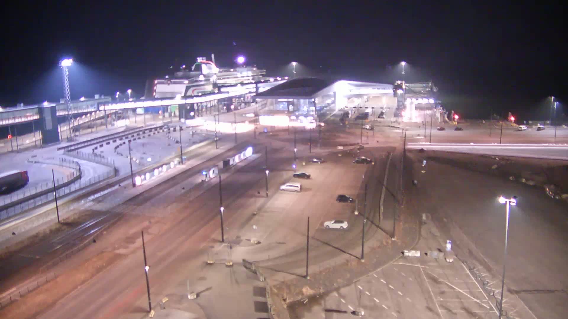 A large ferry is docked at a terminal at night, with several cars parked in the surrounding lots under bright lights.