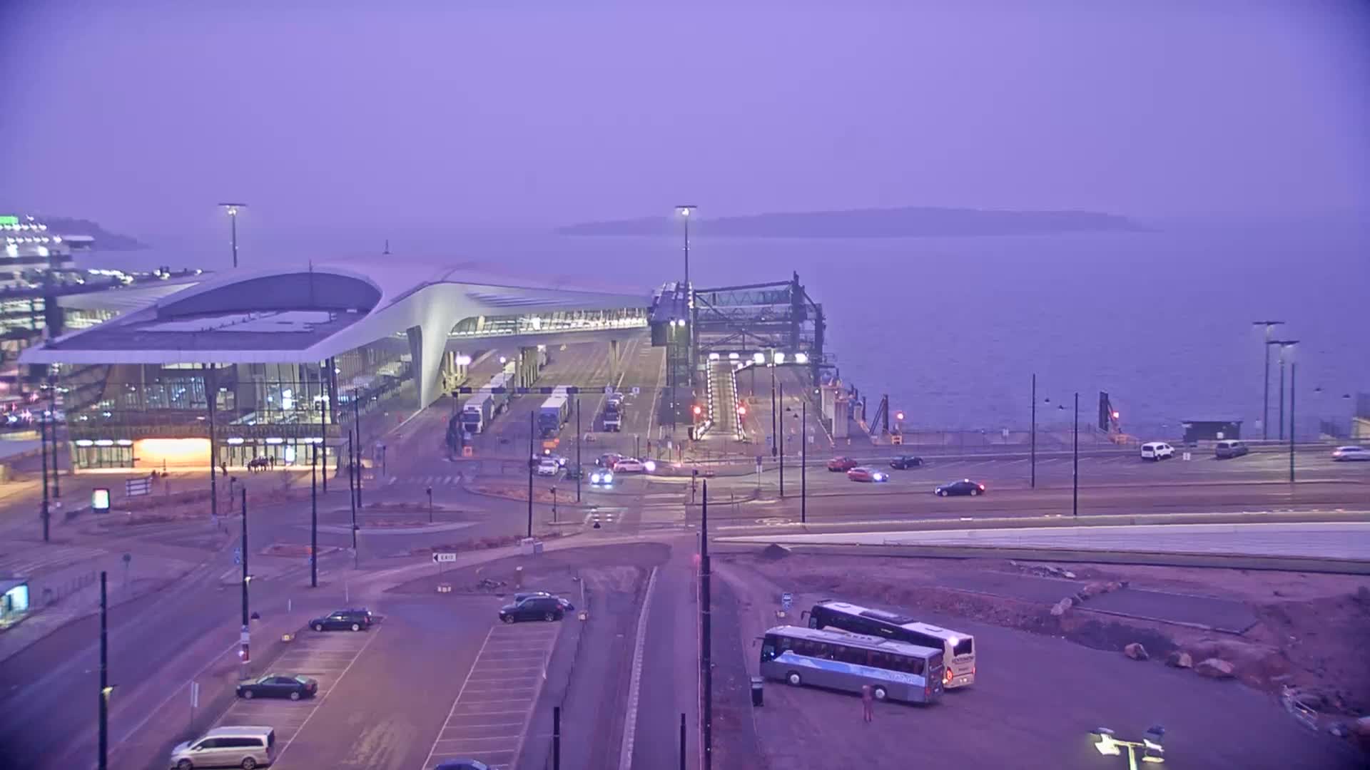 A modern ferry terminal with several vehicles parked nearby sits on the water's edge under a hazy purple twilight sky.