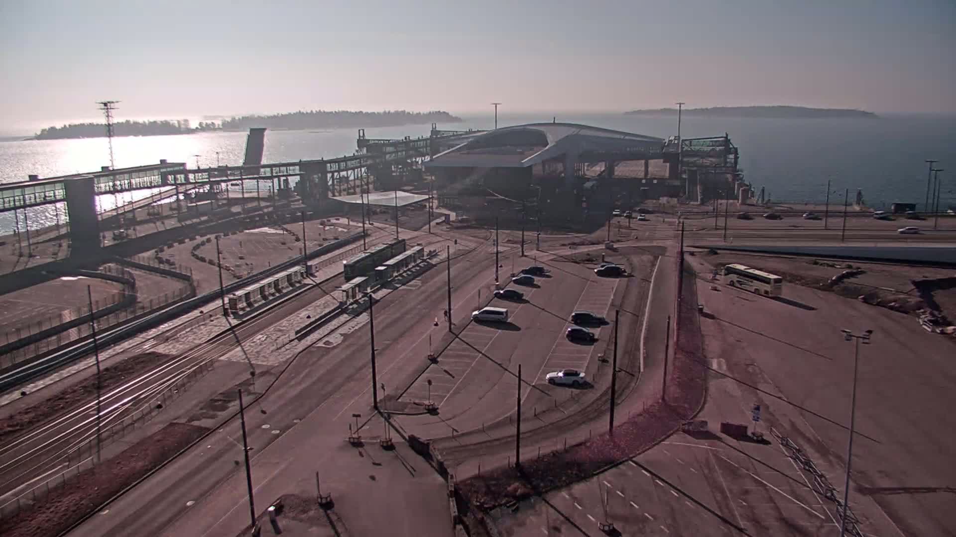 An aerial view shows a ferry terminal with a large building and several vehicles parked nearby on a sunny day.