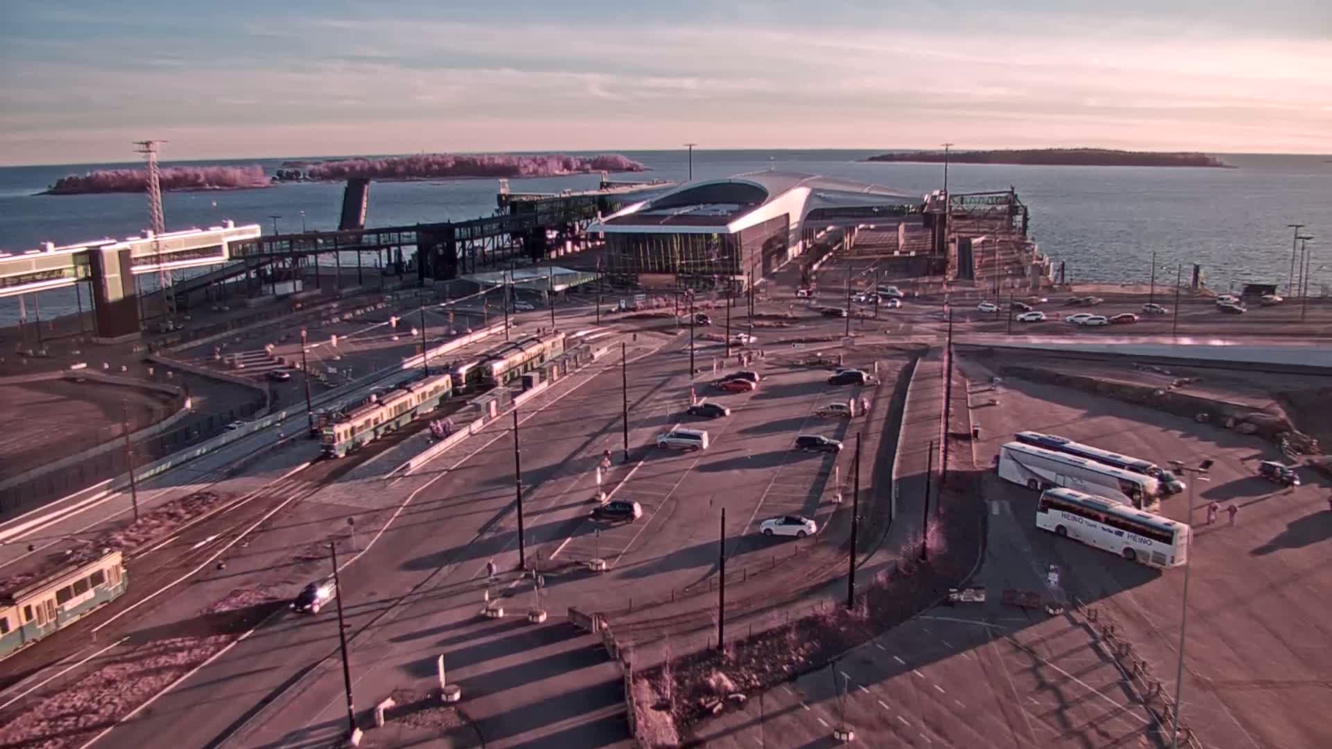 An aerial view of a ferry terminal with trains and buses parked near a modern terminal building on a calm, sunset-lit day.