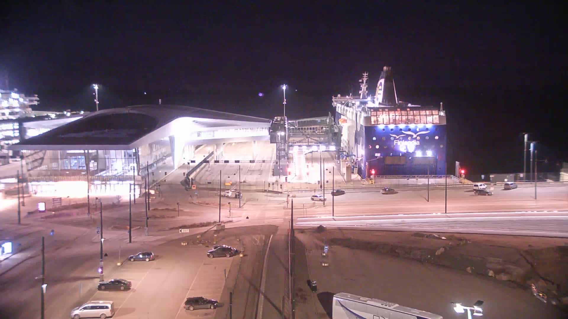 At night, a large ferry is docked at a terminal building with cars parked in the surrounding area.
